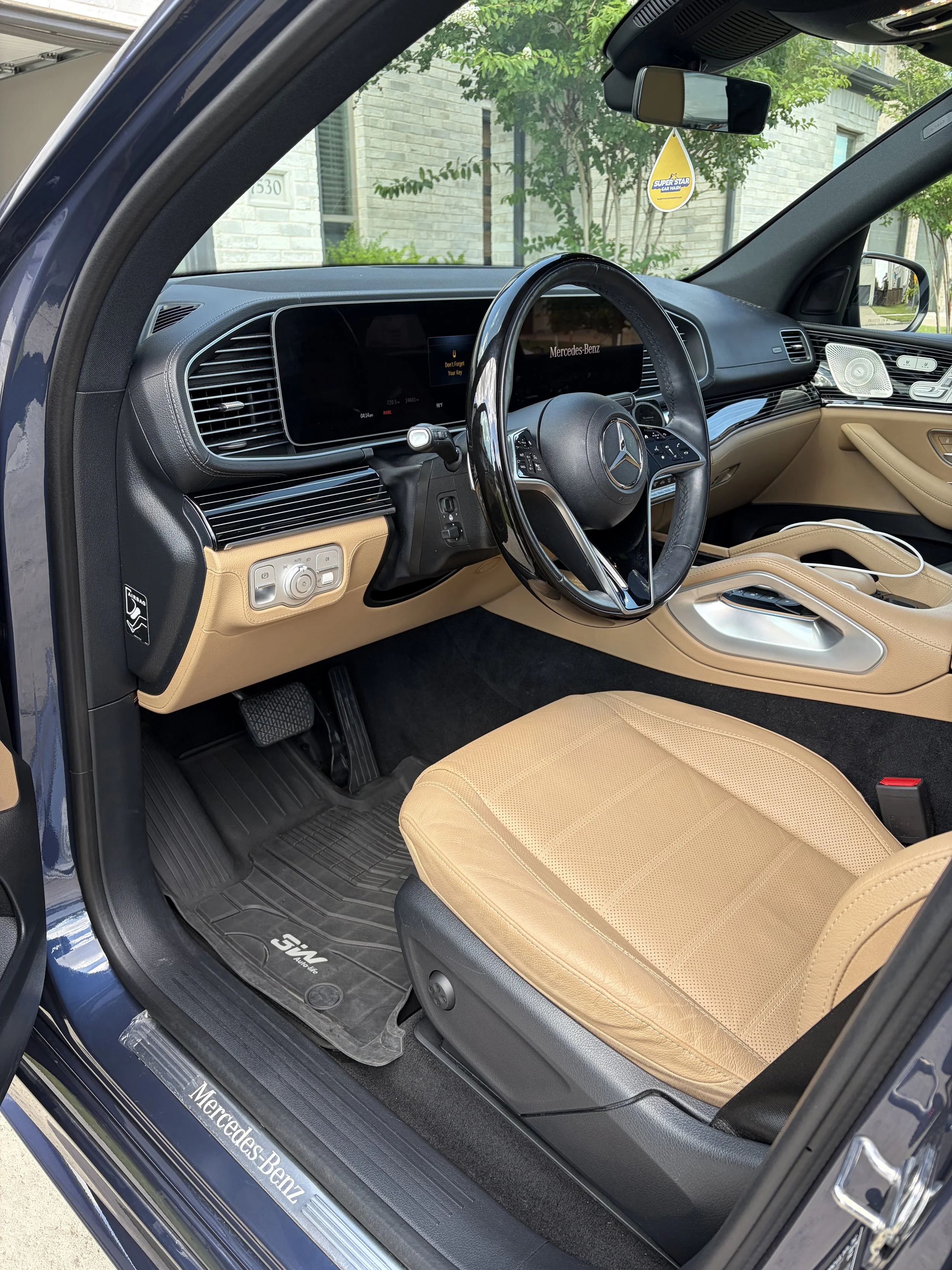 Interior of a Mercedes-Benz vehicle showing a tan leather seat, dashboard with digital displays, a steering wheel with Mercedes-Benz logo, modern controls, and a black and tan color scheme.