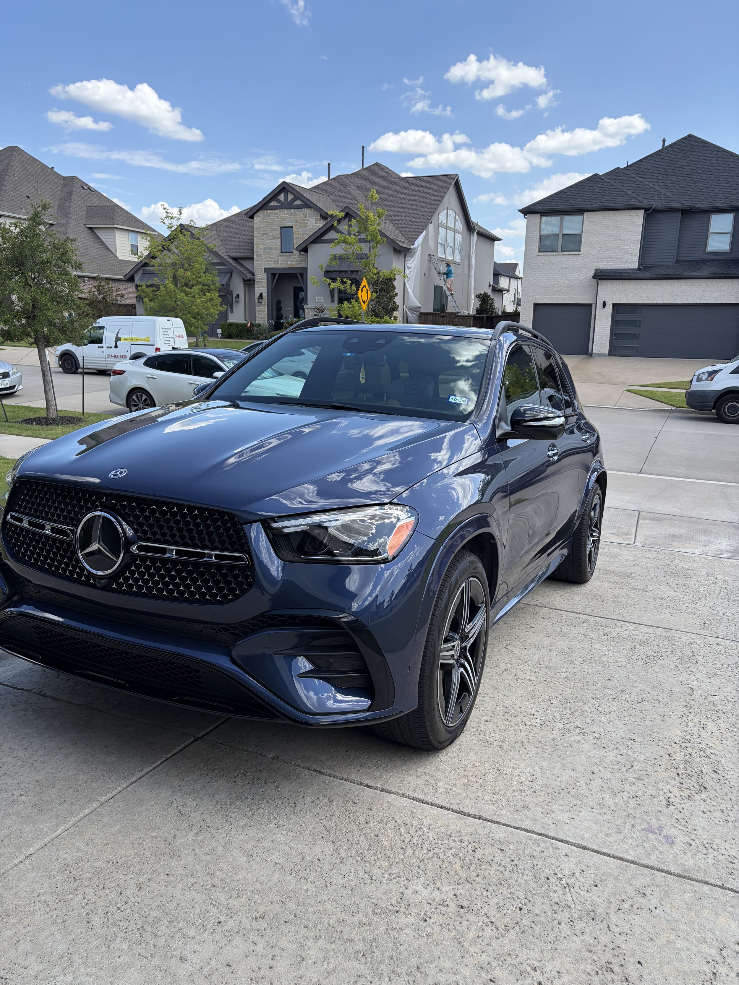 A black Mercedes-Benz SUV parked in a residential driveway with houses and a blue sky with clouds in the background.