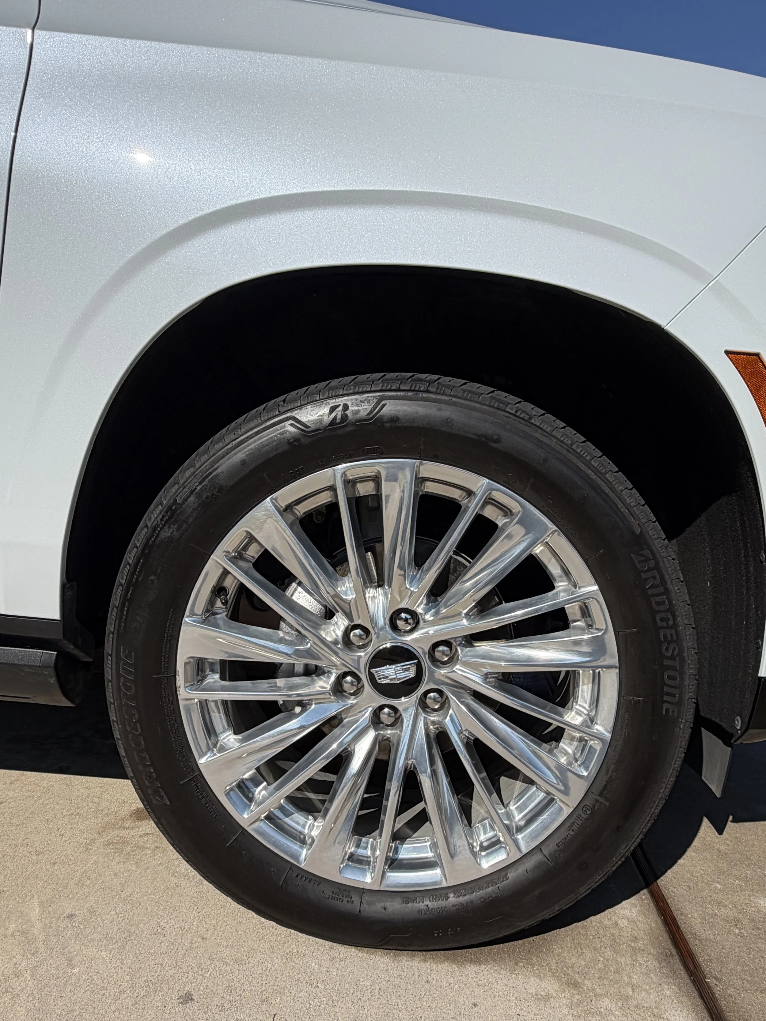 Close-up of a car's front wheel with a silver, multi-spoke rim and a Bridgestone tire, part of the white car body and ground visible.