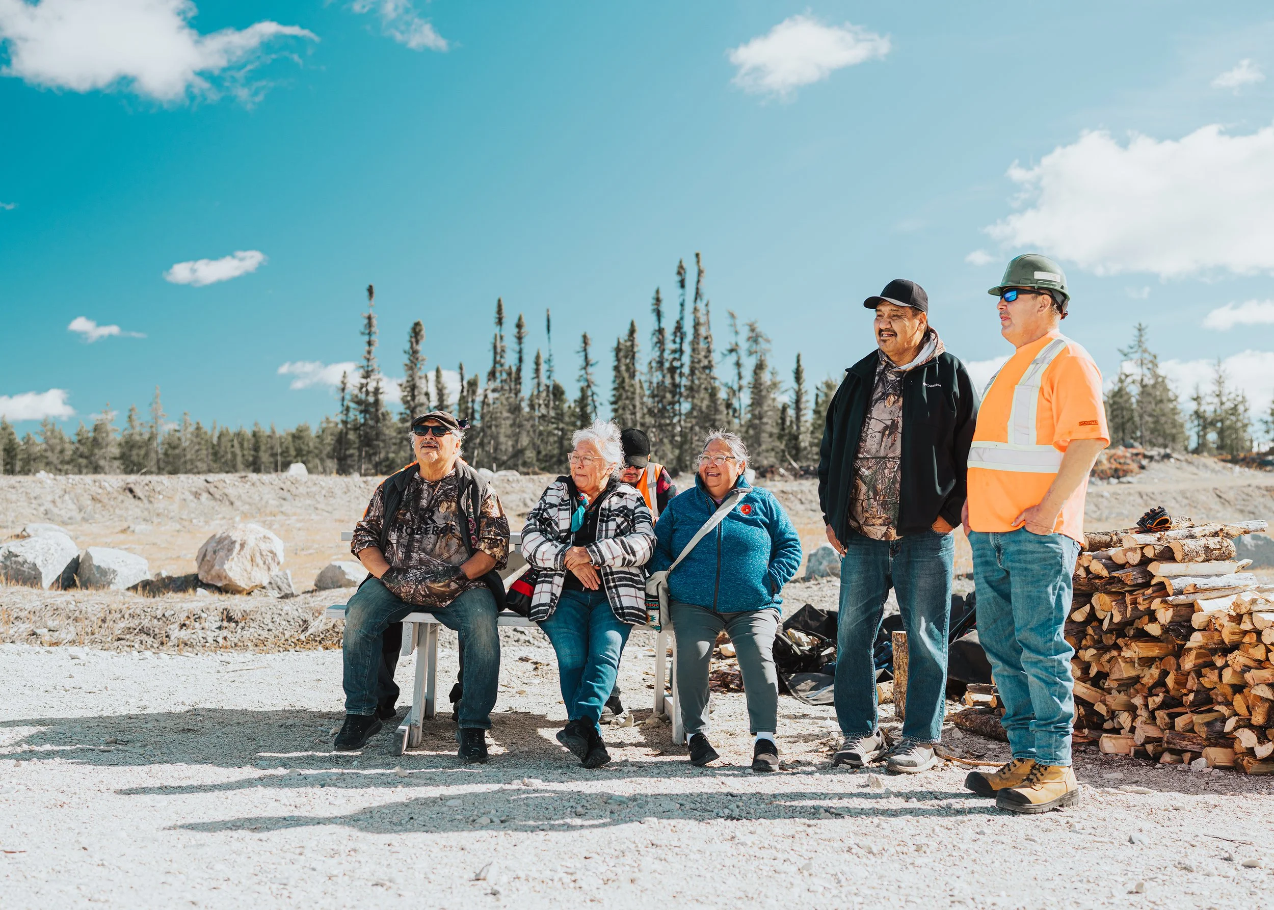 Five community members and workers gathered outdoors at the camp, three seated on a bench