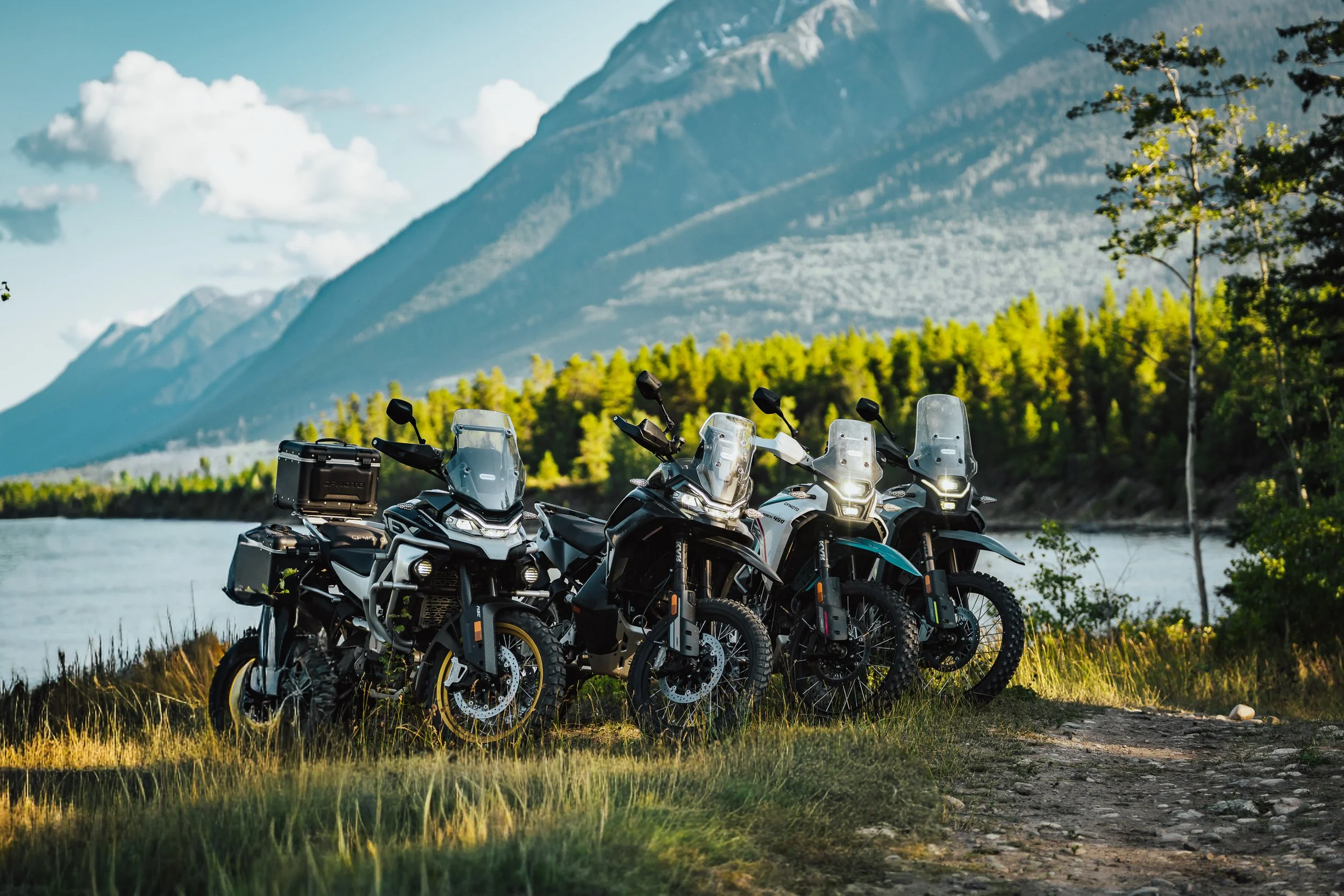 Three CFMOTO IBEX adventure motorcycles parked by a lake under the mountains