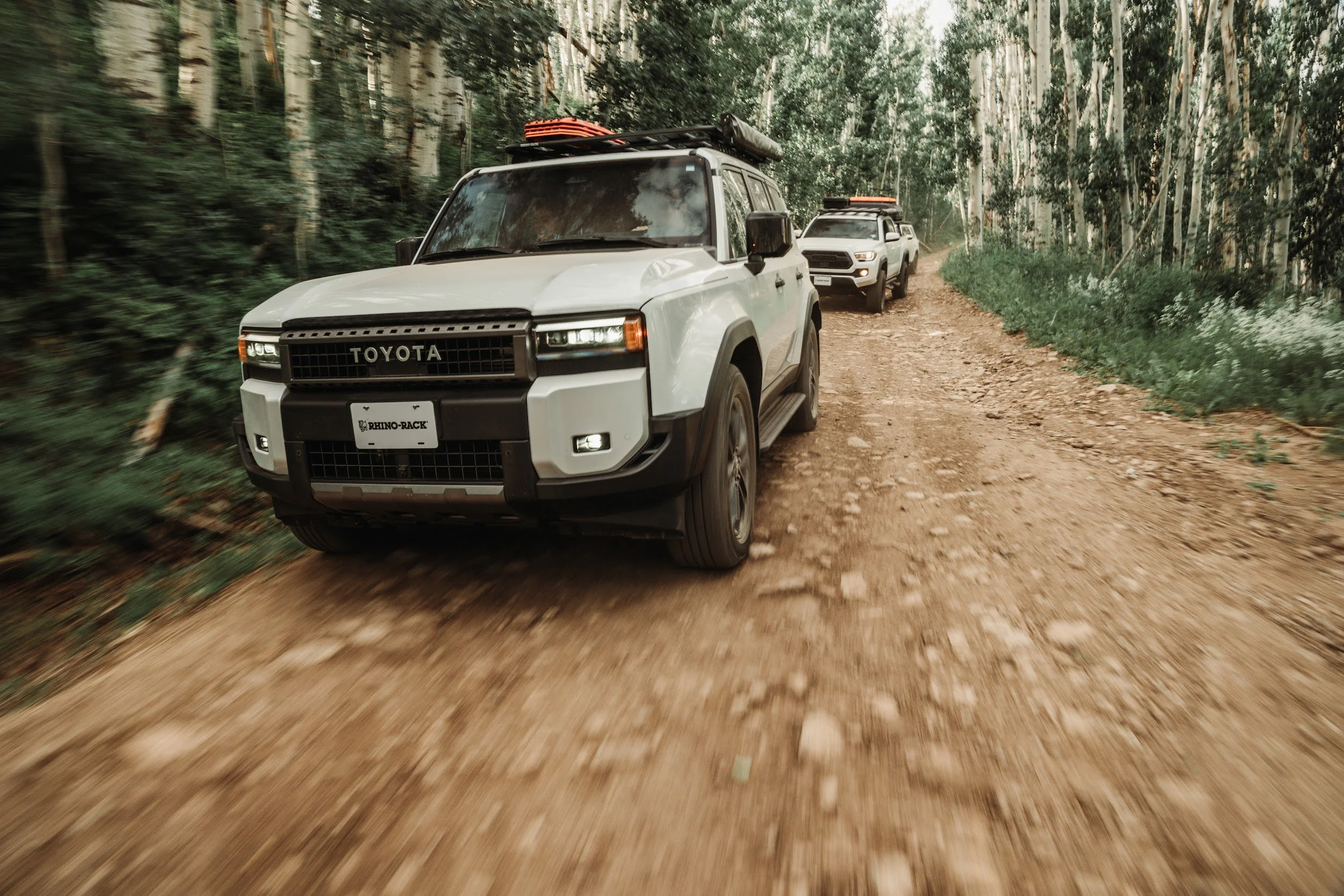Land Cruiser leading the convoy down a dirt trail through aspen trees, shot low with motion blur.