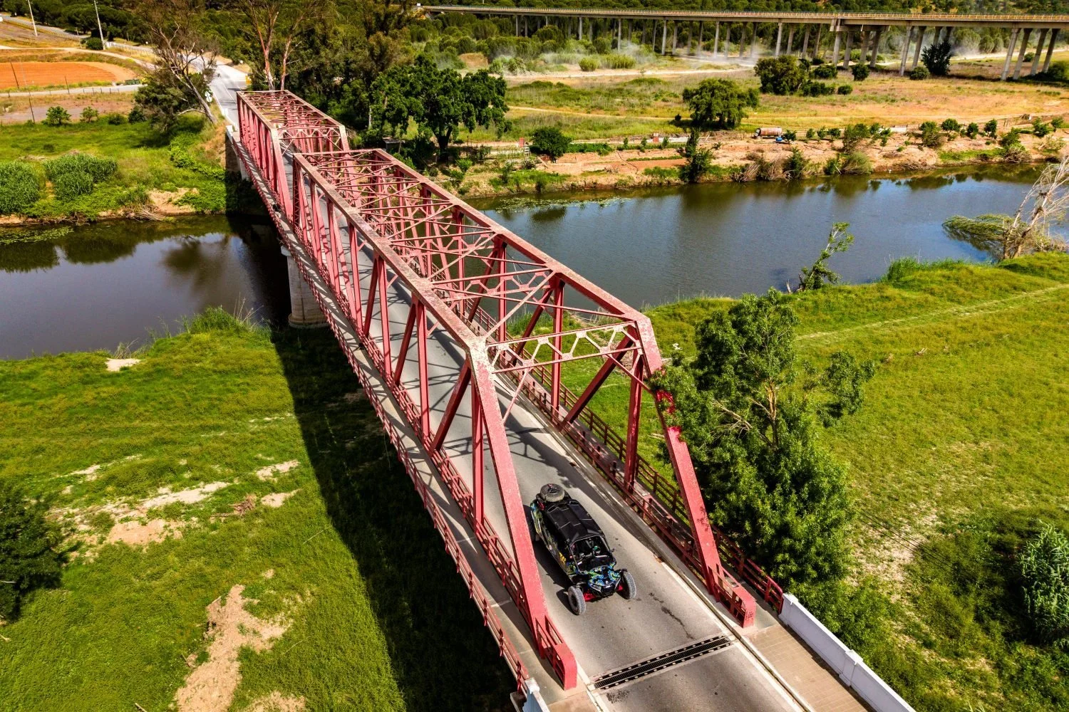 A side-by-side crossing a red iron bridge over a river, seen from above
