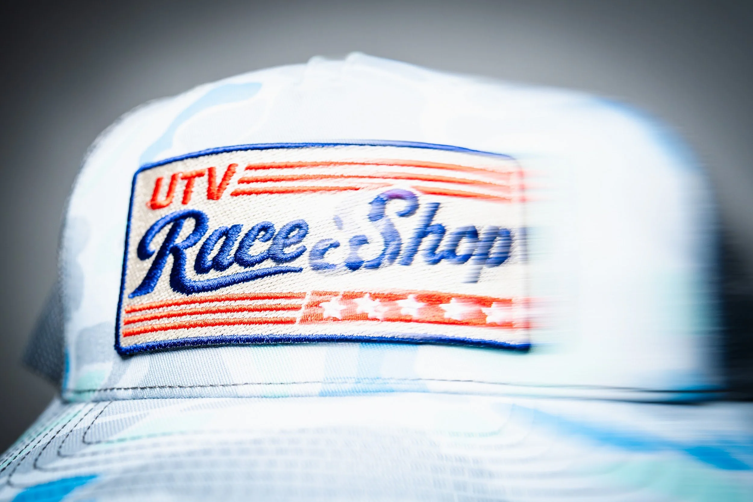 Close-up of a camo trucker hat with an embroidered UTV Race Shop script patch from Capsule 002.