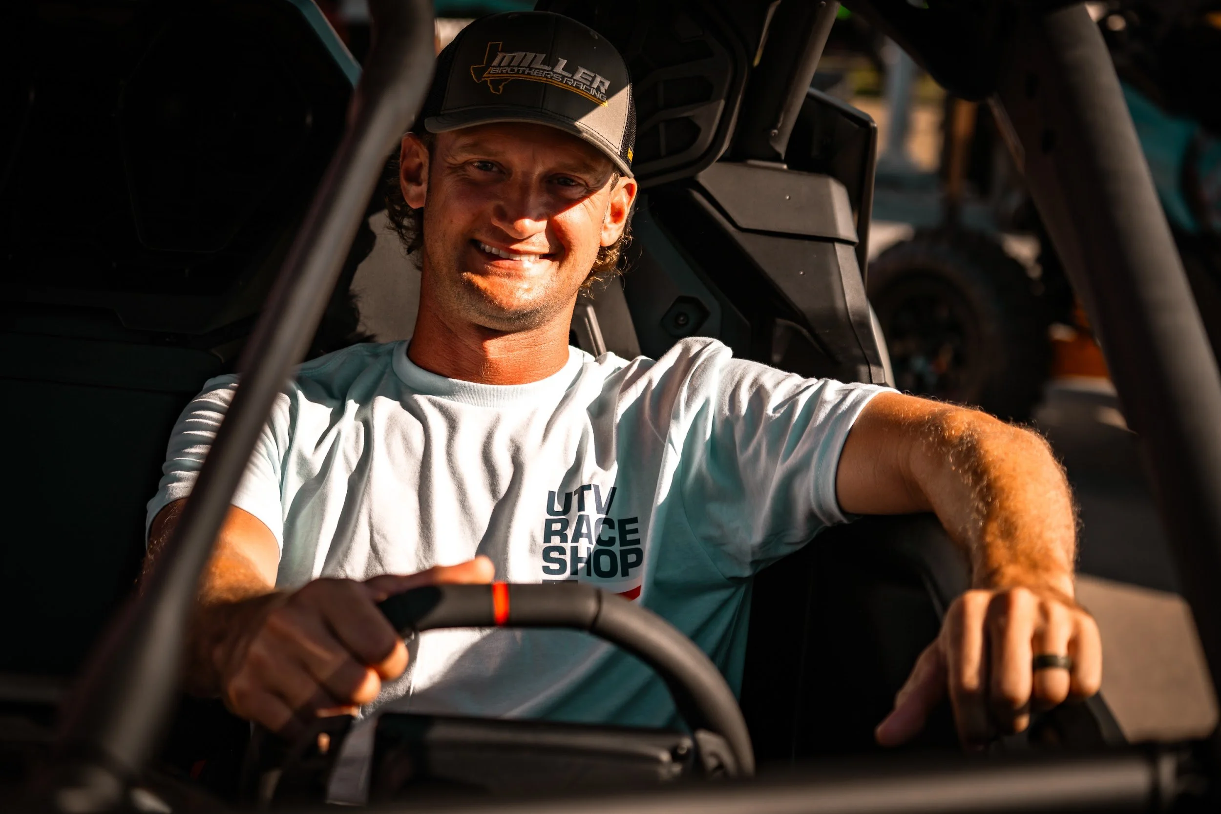 A racer at the wheel of a side-by-side wearing the stacked UTV Race Shop logo tee at the Sand Sports Super Show.