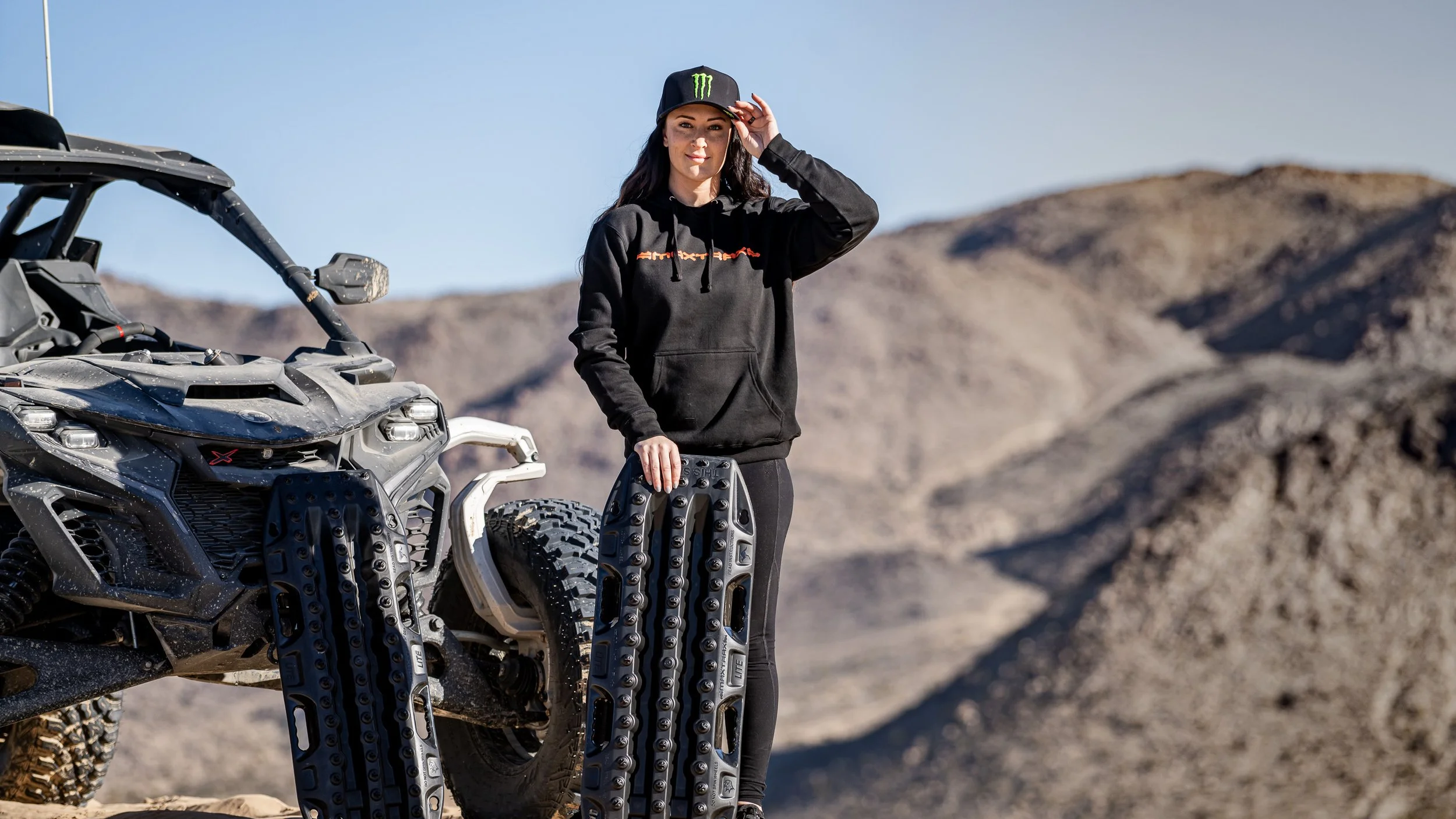 Sara Price beside a Can-Am side-by-side with MAXTRAX recovery boards, desert hills behind