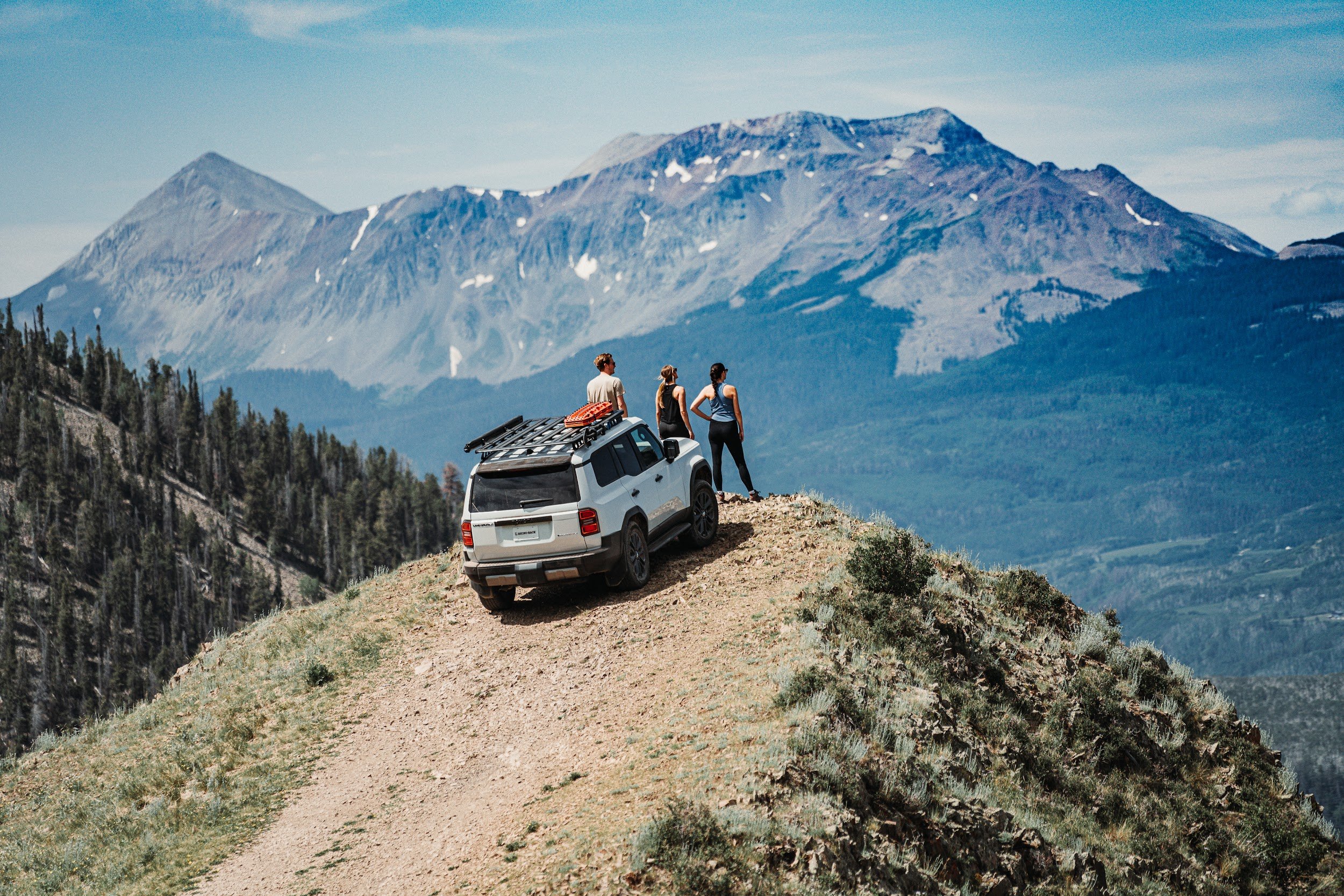 White Toyota parked on a narrow ridge, three people looking out at the peaks across the valley.