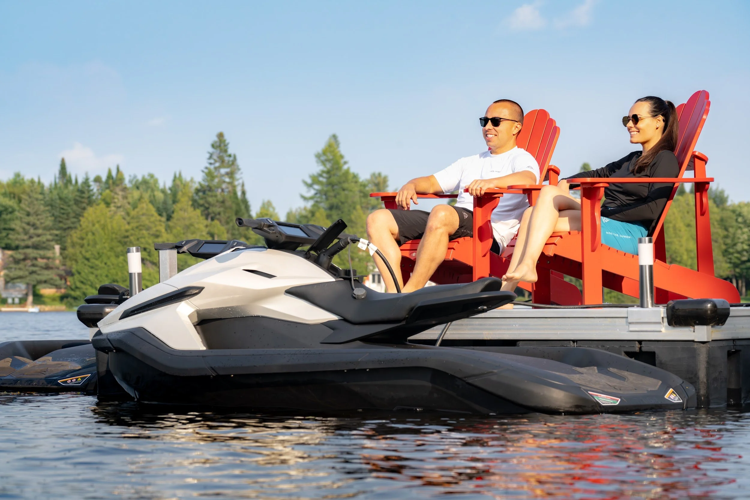 Two people in red Adirondack chairs on a dock, a Taiga Orca moored in front of them