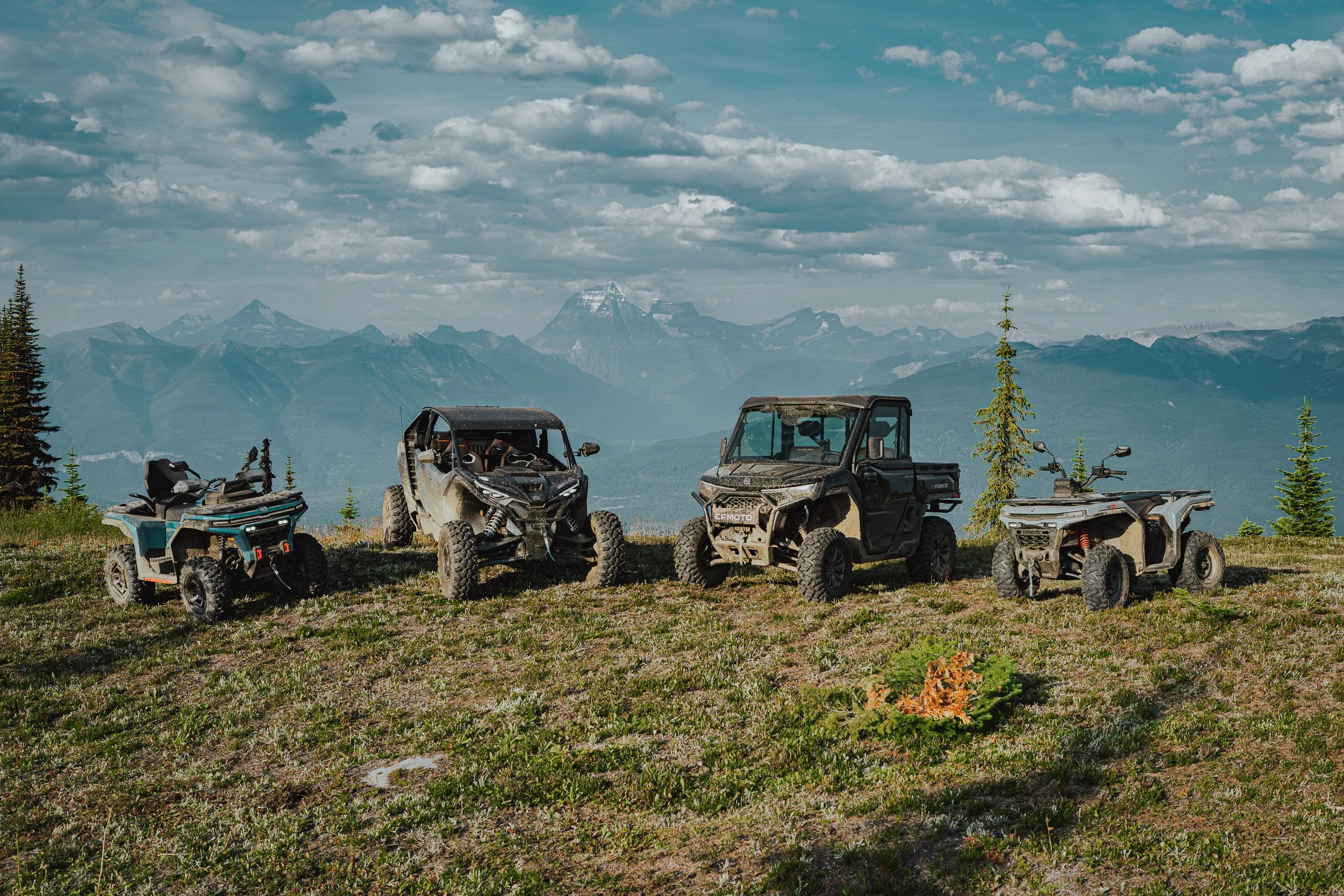 Four CFMOTO ATVs and side-by-sides lined up on an alpine meadow, the Rockies behind