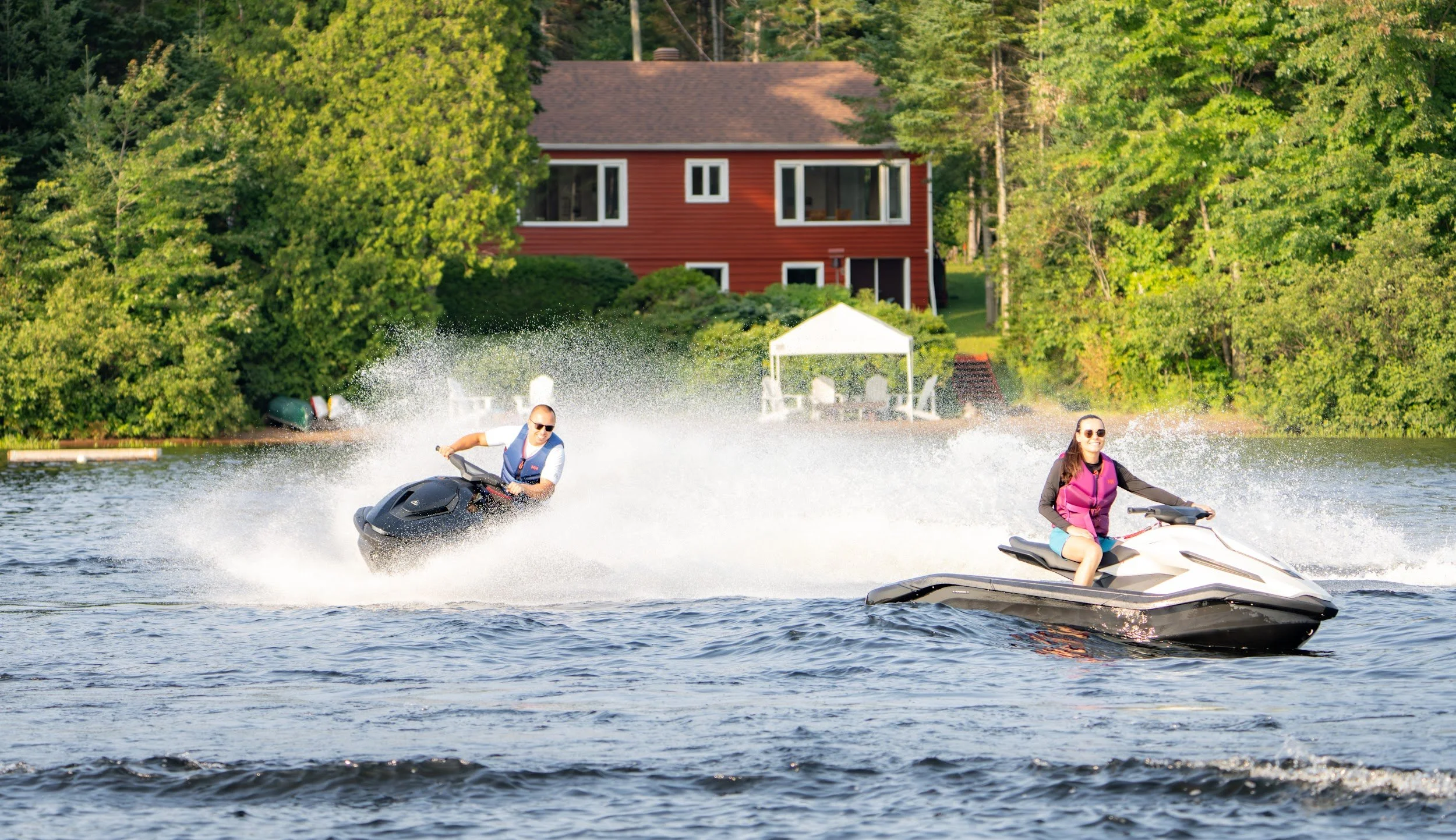 Two riders on Taiga Orca Performance watercraft throwing spray in front of a red lakeside cottage