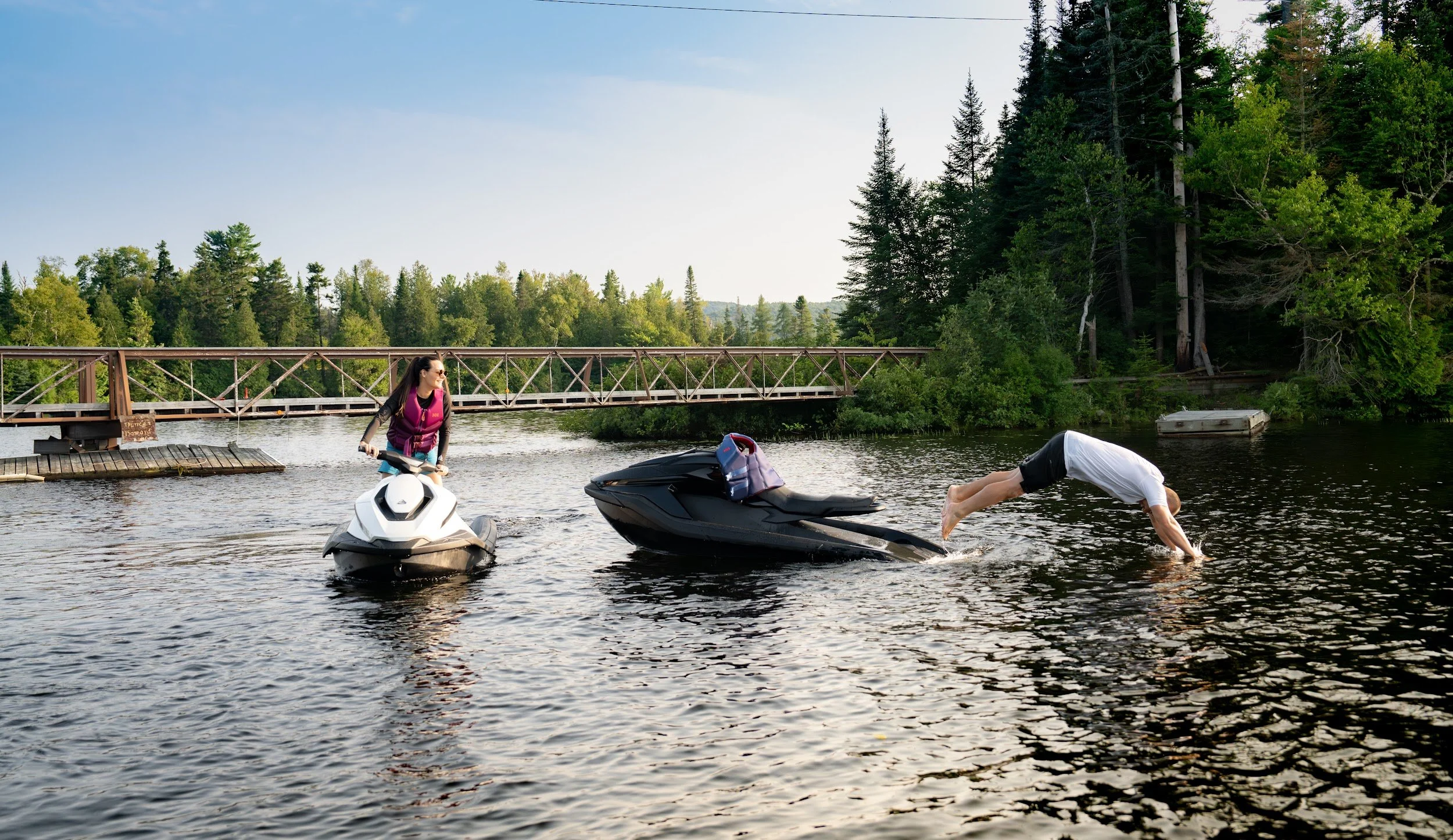 Rider diving off a Taiga Orca into the lake near a footbridge while another rider watches