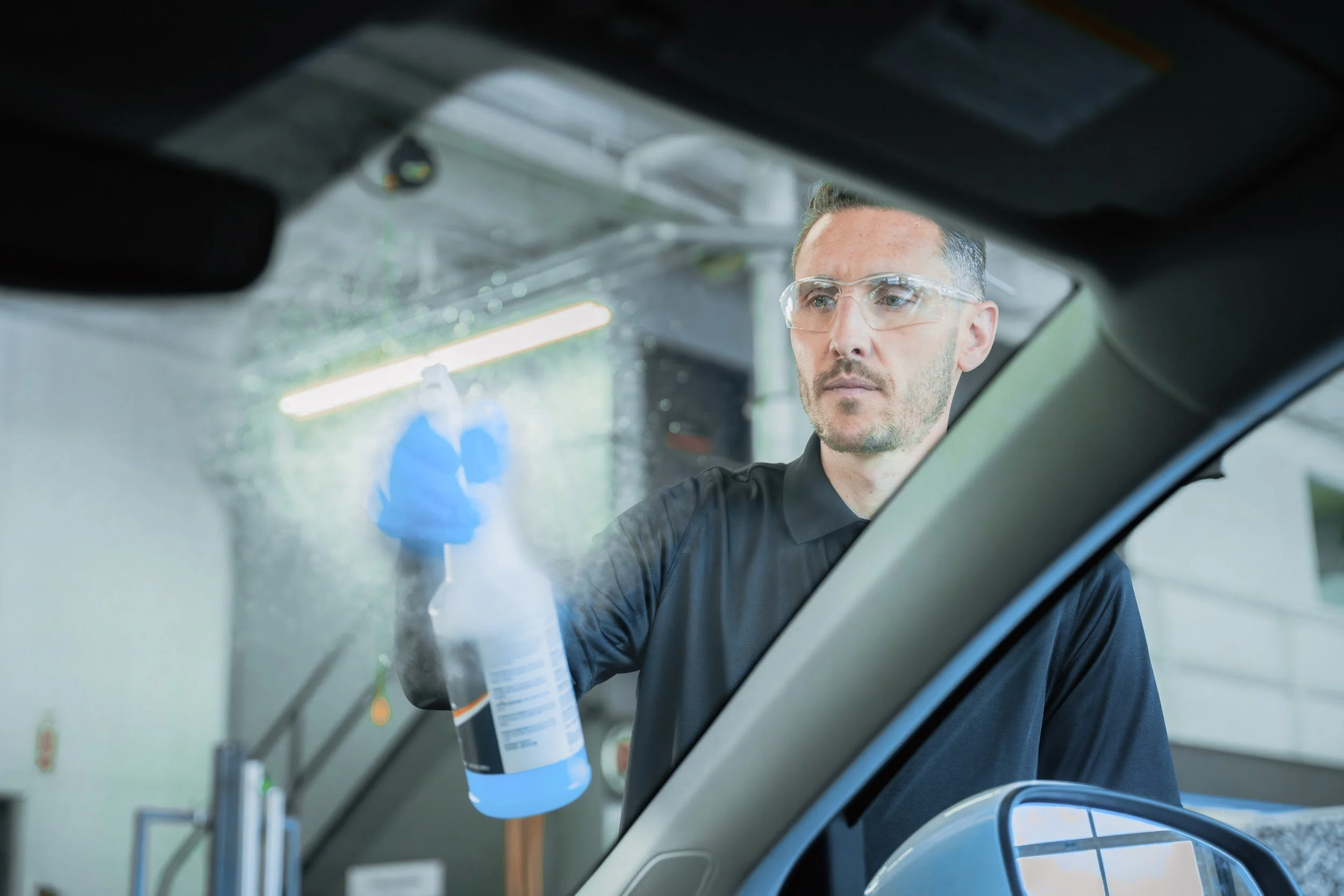 Technician spraying rain repellent on a windshield, seen from inside the car