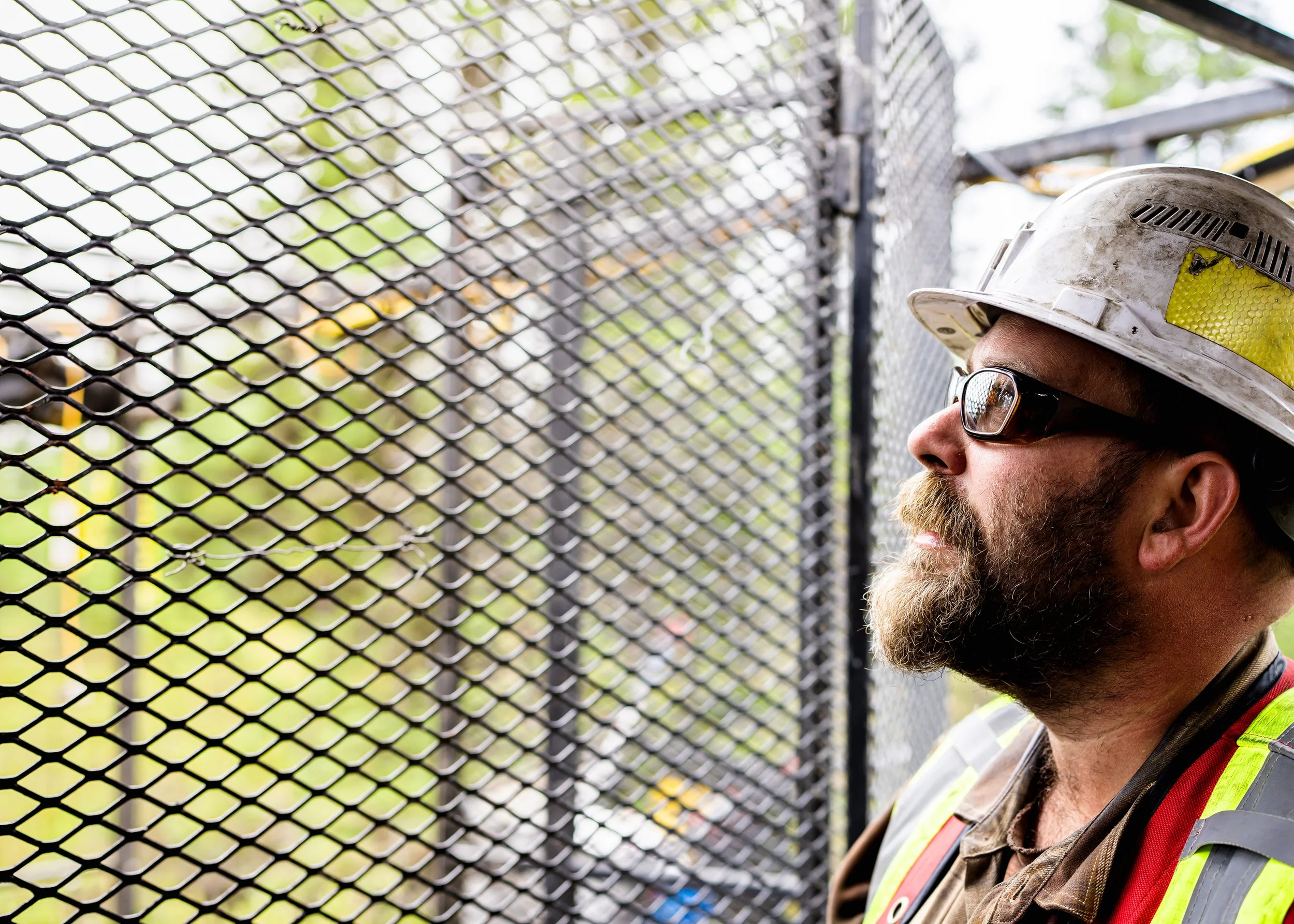 Bearded worker in a hard hat and safety glasses looking through a steel mesh gate