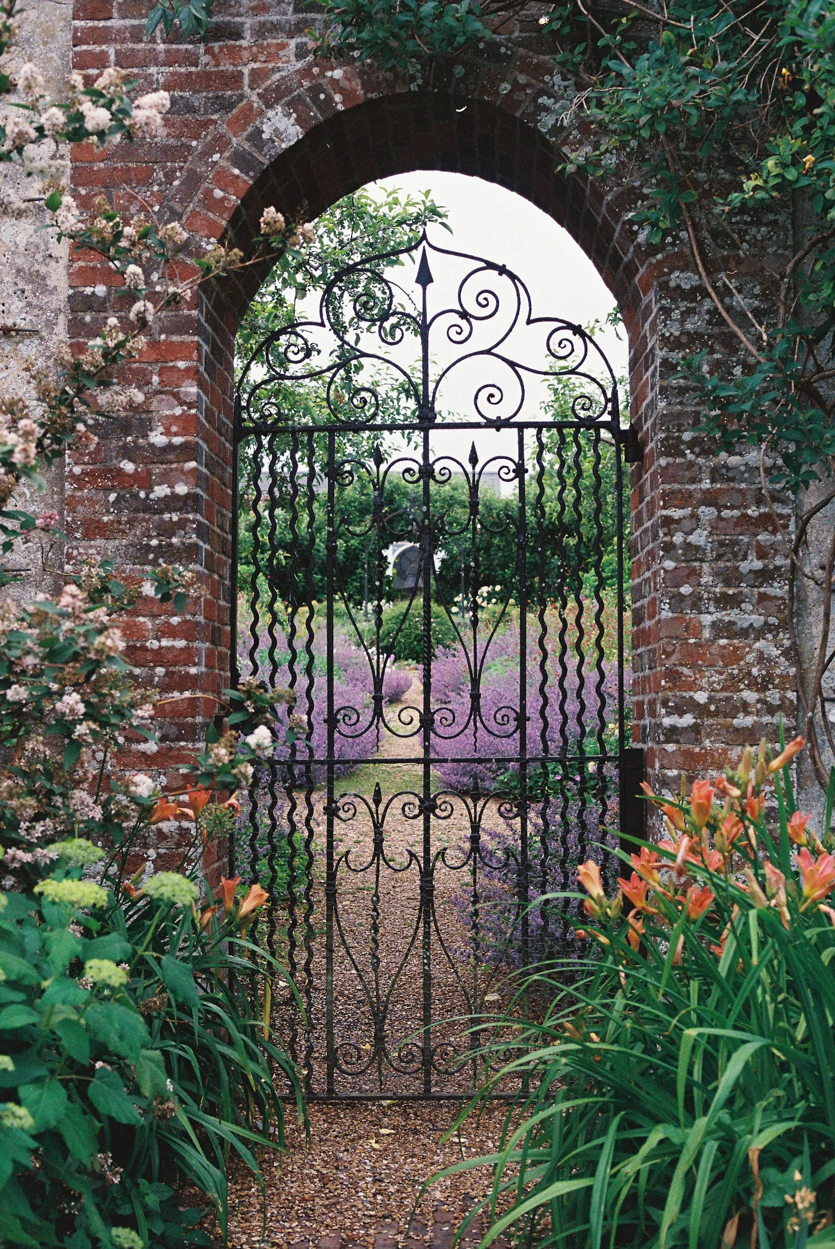 Wrought-iron garden gate set in a red brick archway, surrounded by flowers and greenery.