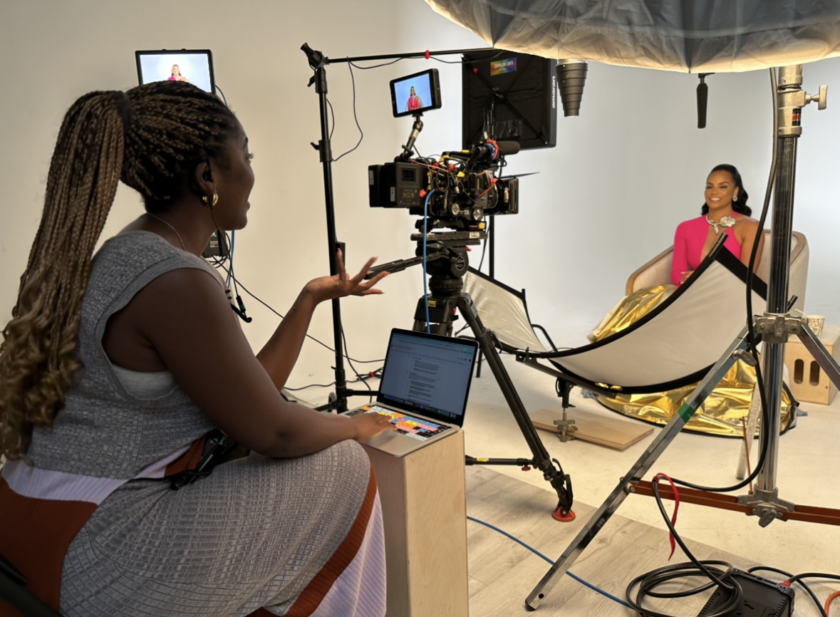 A woman director on a video set filming a woman in a pink dress with gold accents, while a crew member manages camera equipment and lighting in a studio with a white background.