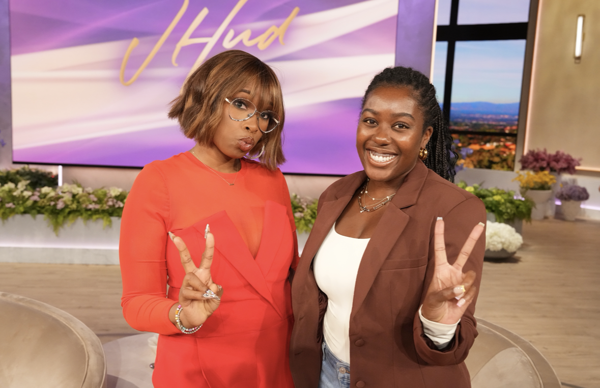 Two women posing on a TV show set, with a purple and yellow background, floral decorations, and a cityscape view in the window, both making peace signs and smiling.