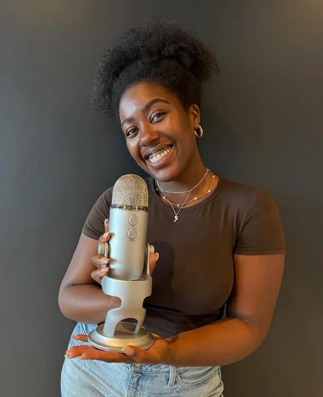 A smiling woman with natural curly hair holding a large silver microphone against a dark background.