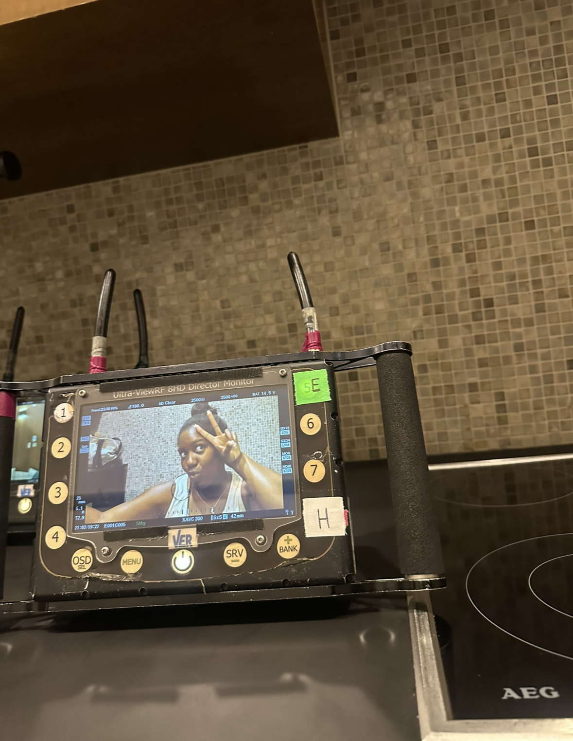 Close-up of a camera monitor showing a woman making a peace sign near her eye, sitting at a table with a mosaic tile wall in the background.