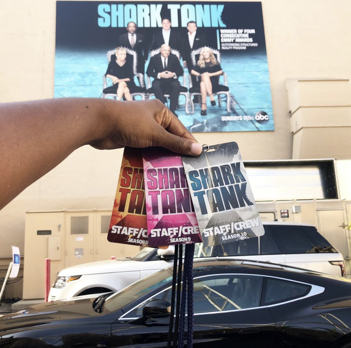 Hand holding three badges for the show 'Shark Tank', each with different colors, in front of a parking lot with a black car and a white vehicle. A large promotional poster for 'Shark Tank' is on the building wall in the background.
