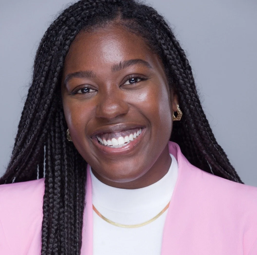 A smiling woman with braided hair, wearing a pink blazer and gold earrings, against a neutral background.