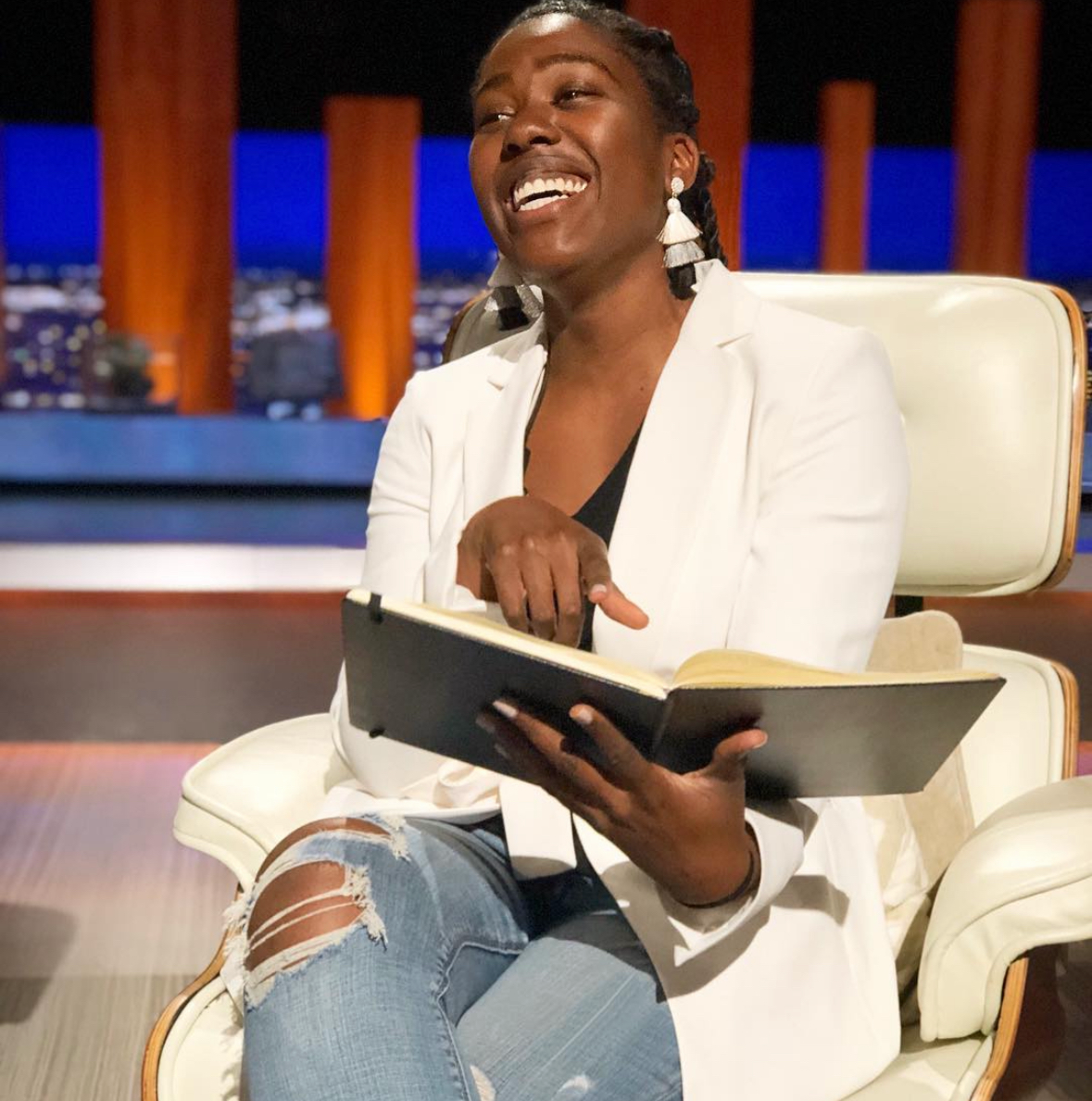 A woman with braids and earrings, wearing a white blazer and ripped jeans, smiling, sitting in a cream-colored chair, holding an open notebook or book.