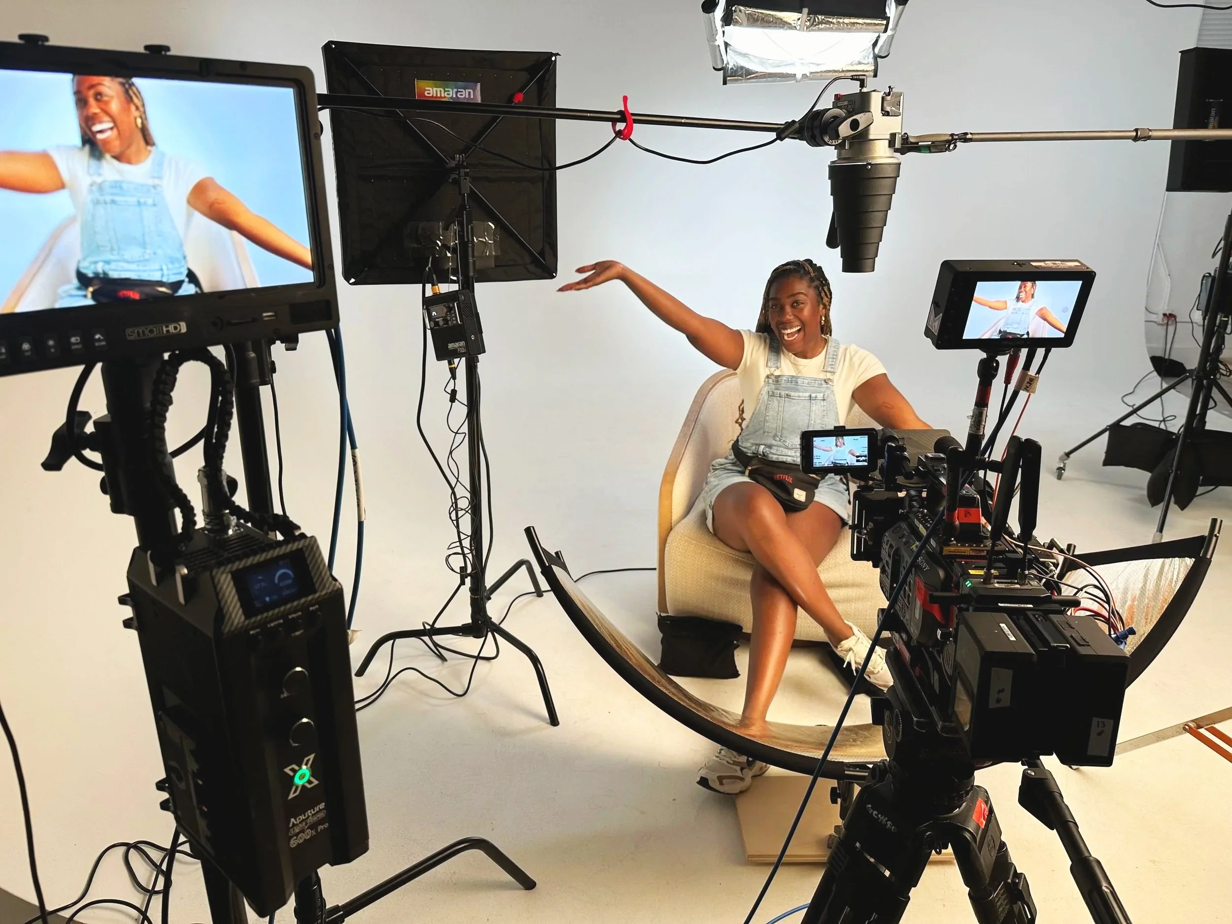 A woman with braided hair, wearing a yellow shirt and denim overalls, smiling and sitting on a chair with her legs crossed, in front of a camera setup in a studio. Multiple cameras and monitors are capturing her pose and facial expression.