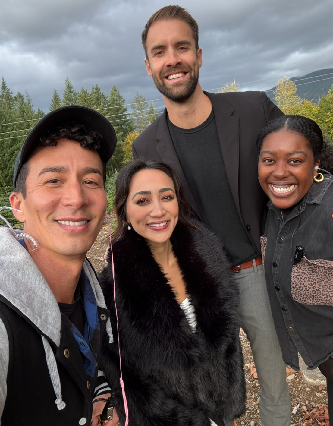 Group of four friends smiling outdoors with trees and cloudy sky in the background.
