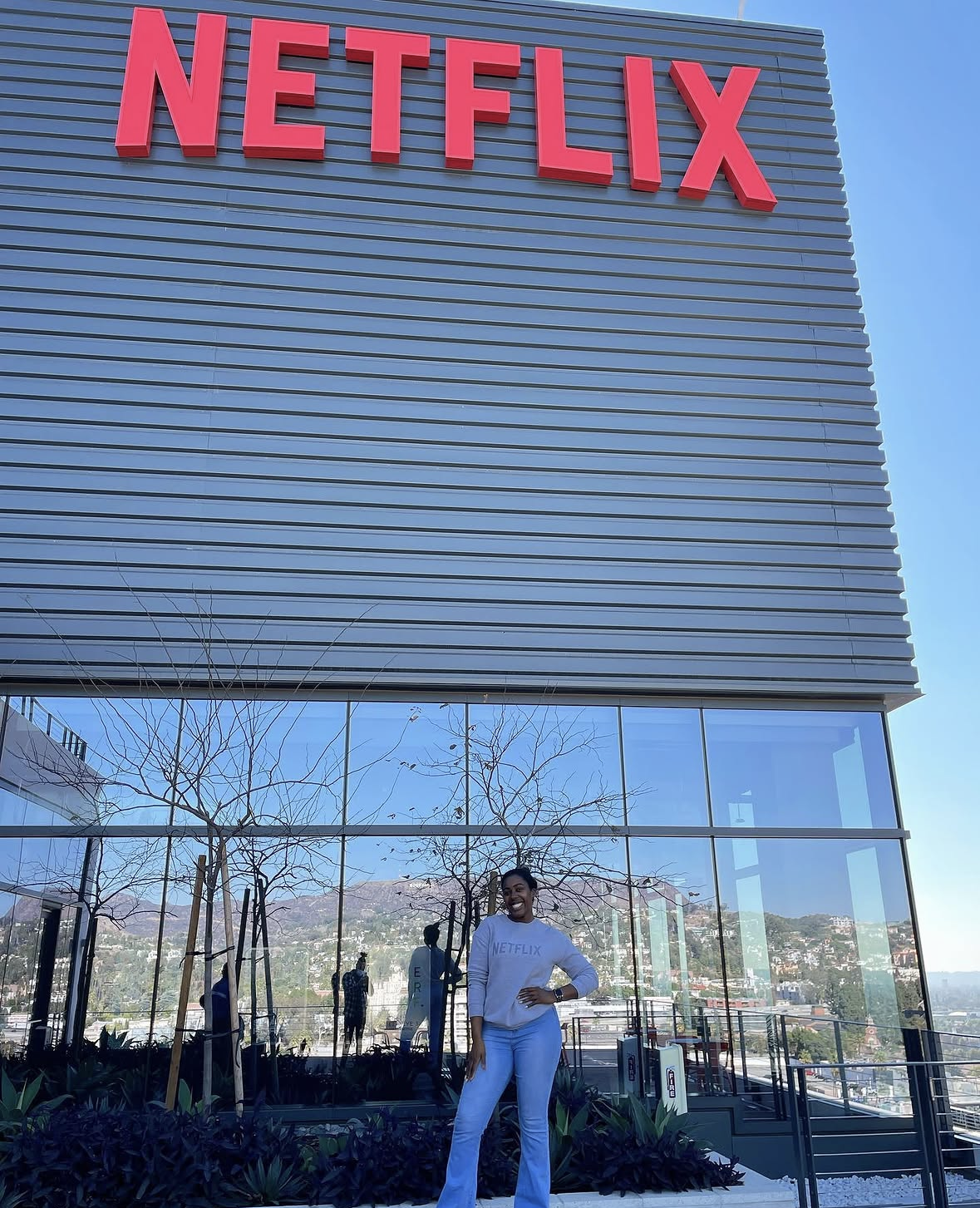 A woman smiling and laughing in front of a large Netflix sign on a modern building with glass windows and a city view in the background.
