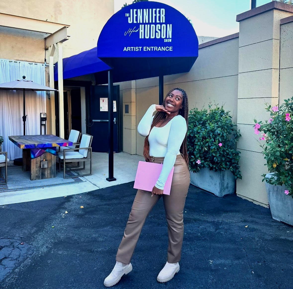 Woman smiling and posing outside the Jennifer Hudson Show artist entrance, holding a pink folder, wearing a white top, beige pants, and white boots with a blue canopy and potted plants in the background.