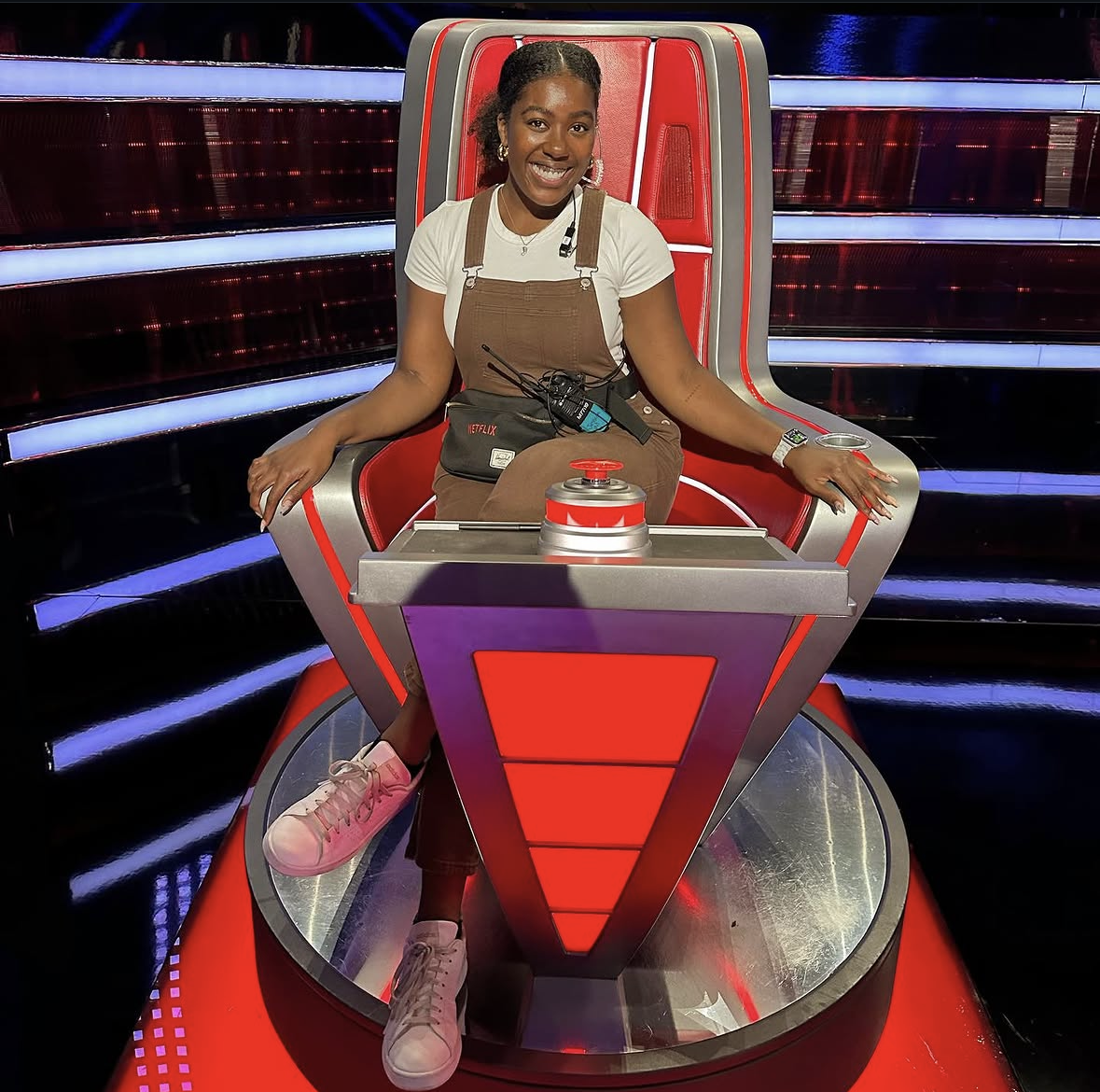 A woman sitting in the chair from The Voice with red, white, and gray colors, smiling, wearing a white t-shirt, brown overalls, and pink shoes.