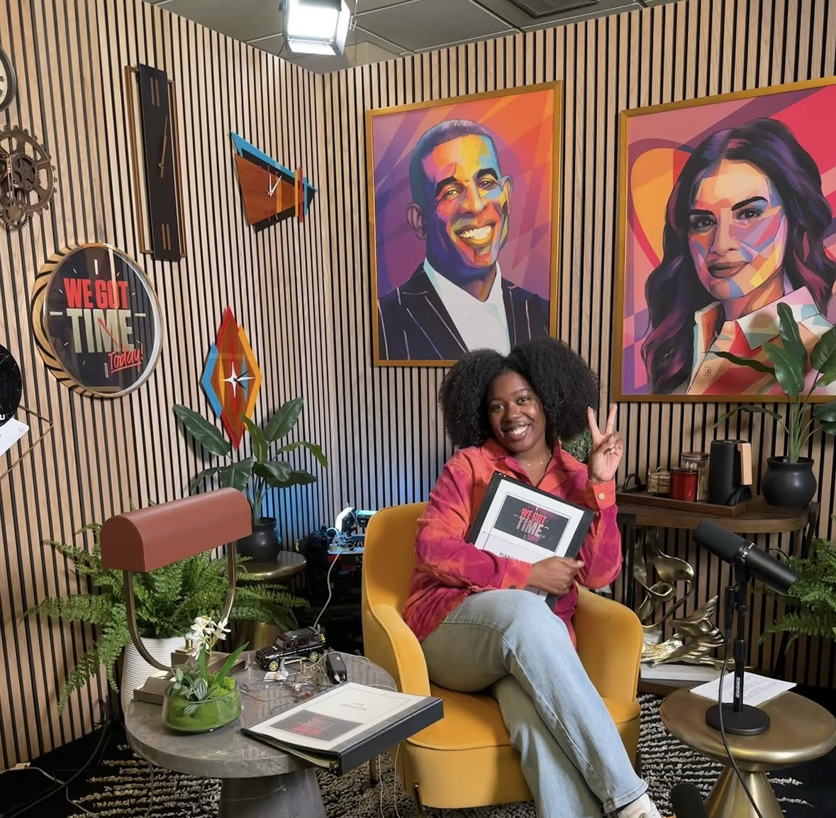 A woman with curly hair is sitting in a yellow armchair, smiling and making a peace sign. She is holding a framed certificate. The room has wooden paneling, colorful portraits on the wall, plants, a microphone on a small table, and various decorative items.