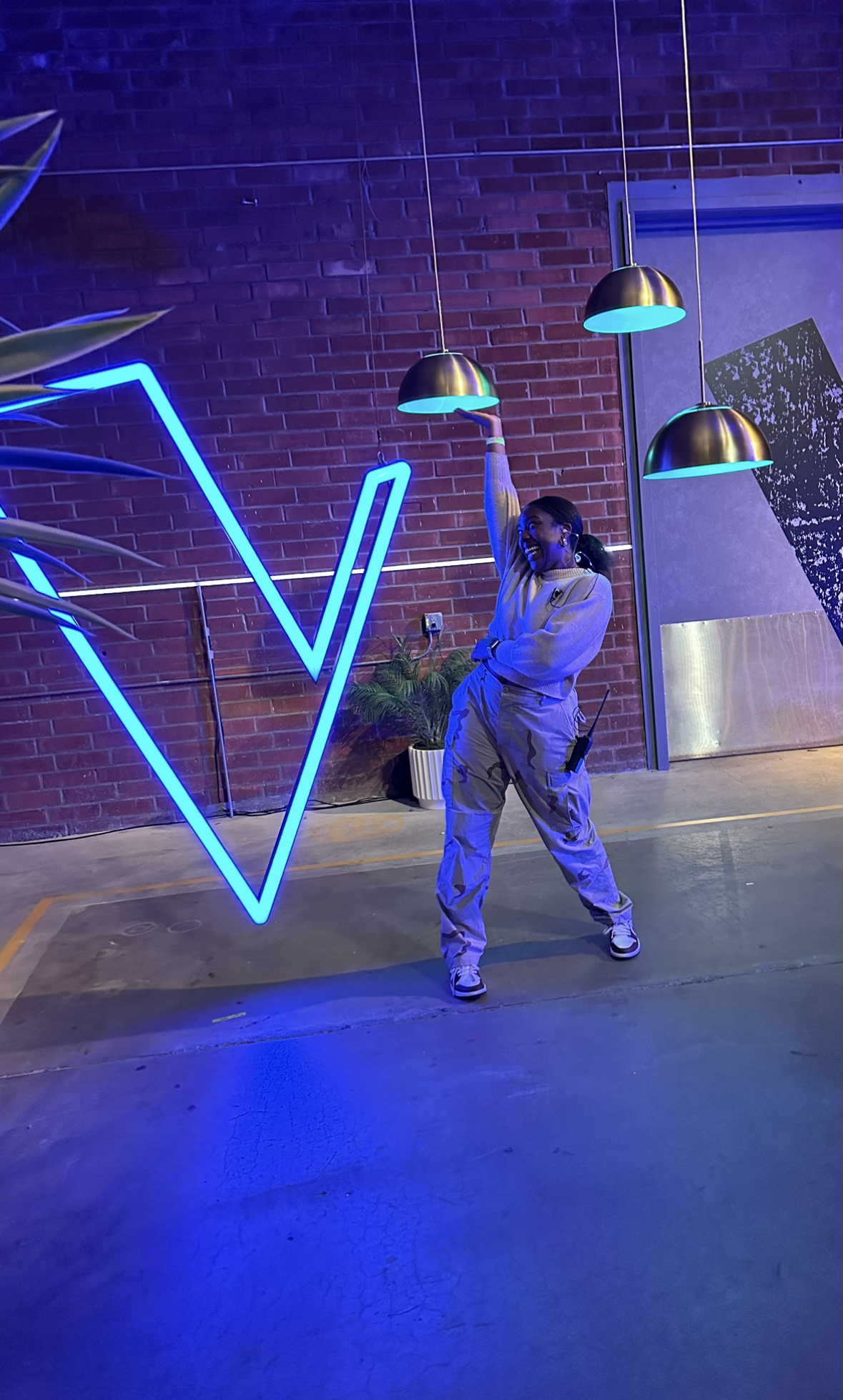 A woman standing in a parking garage with brick walls, brightly lit by hanging black and blue lights, smiling and posing with one arm raised.