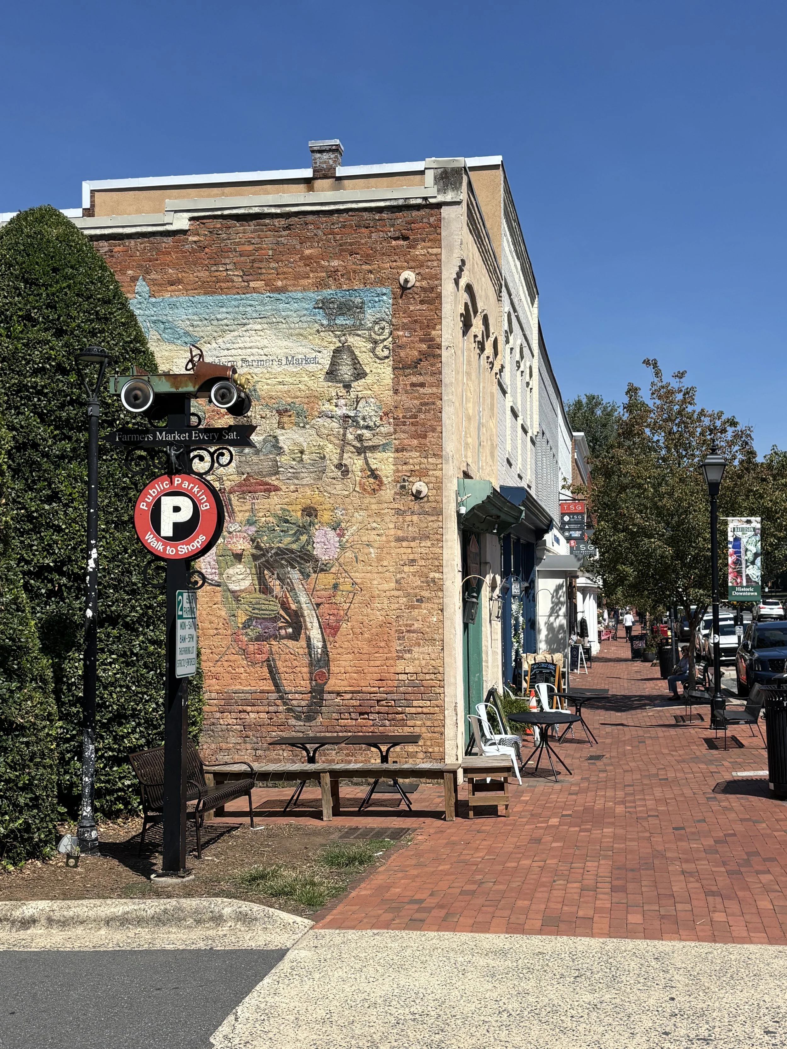 A street scene with outdoor seating, brick sidewalk, a mural on a brick building wall, and trees under a blue sky.