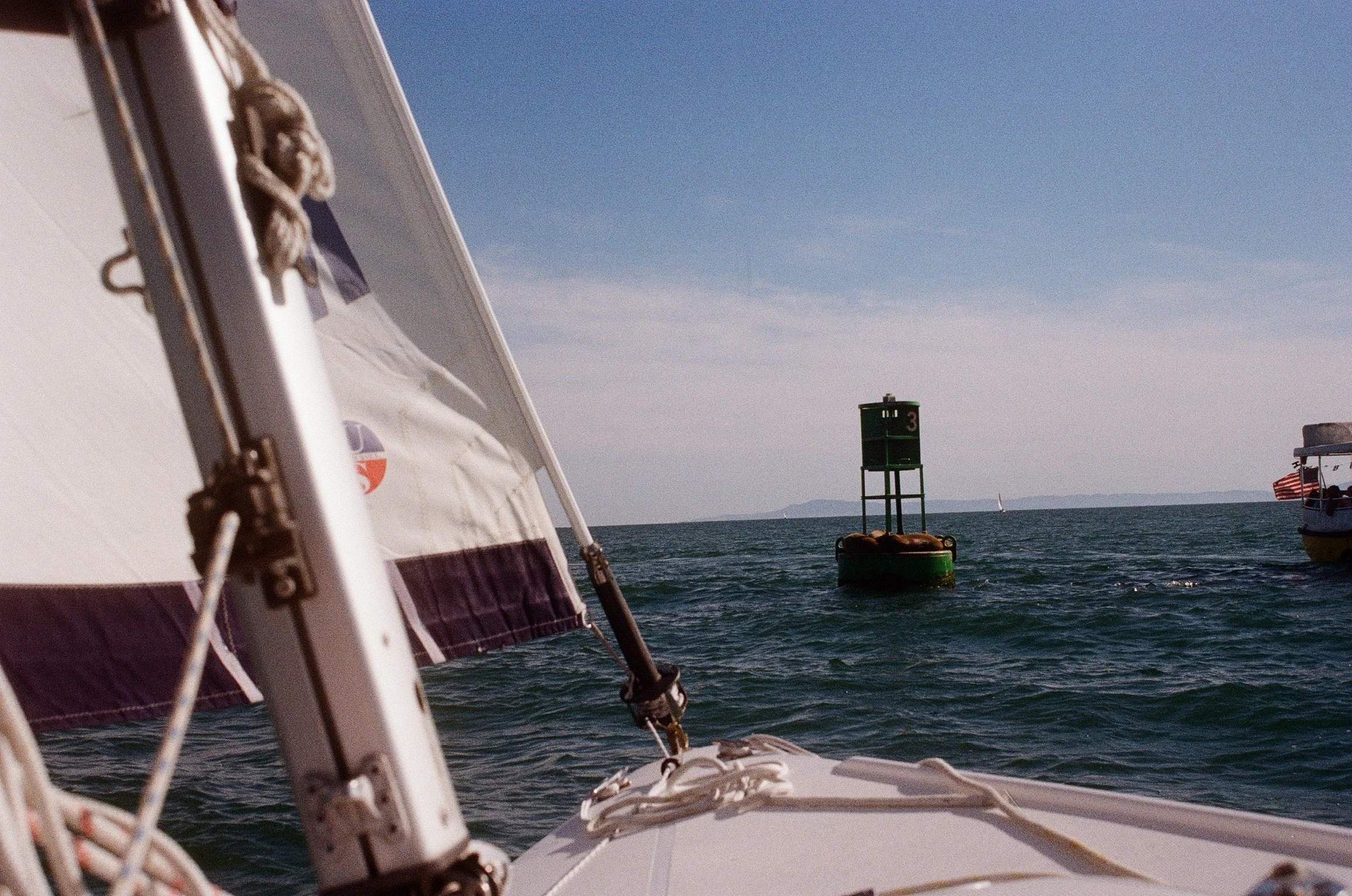 View from a boat capturing nautical elements at sea, including a buoy, a lighthouse, and sailboats in the background under a clear blue sky.
