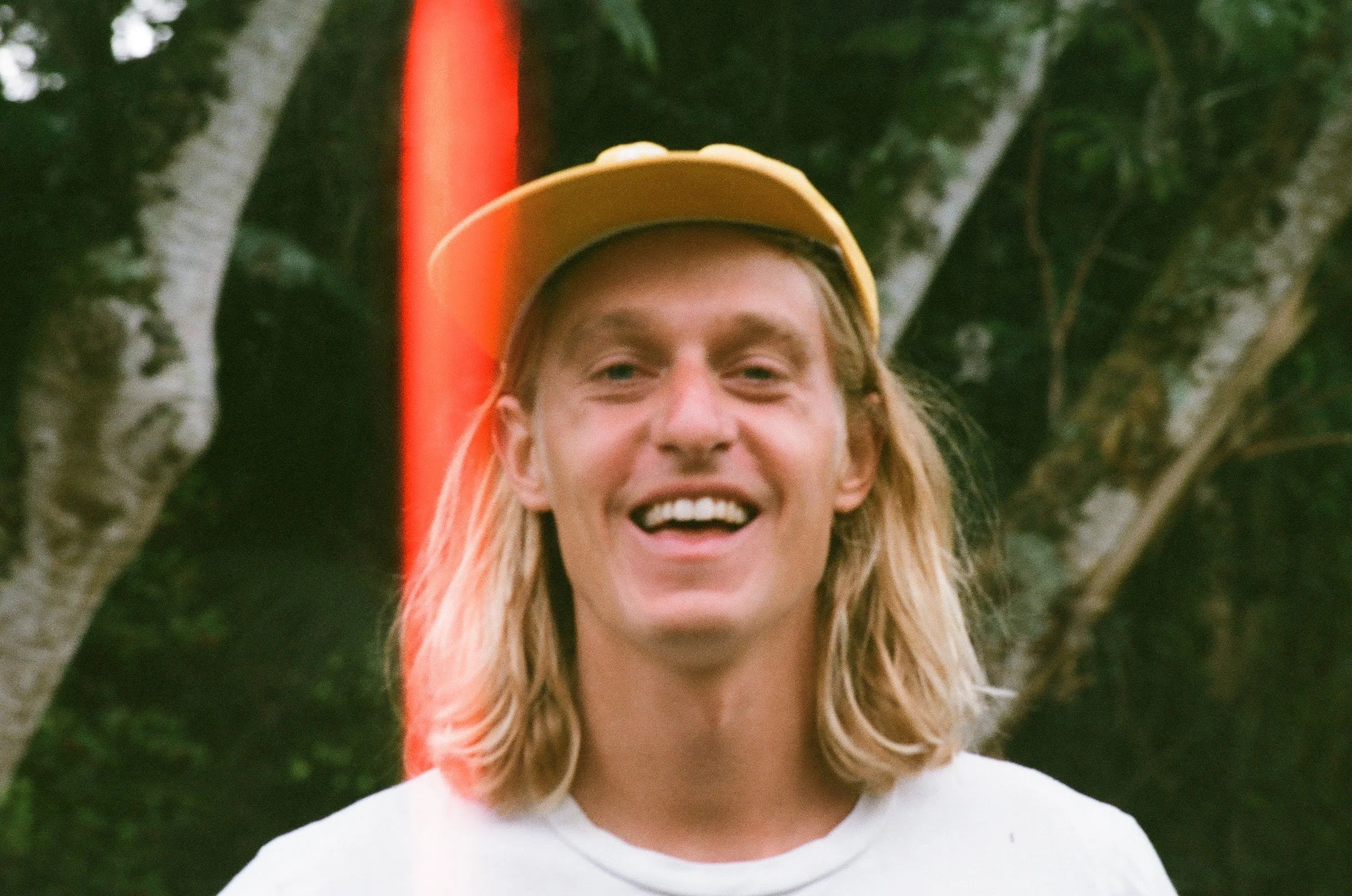 A woman with blonde hair wearing a yellow hat and a white shirt, smiling outdoors among trees.