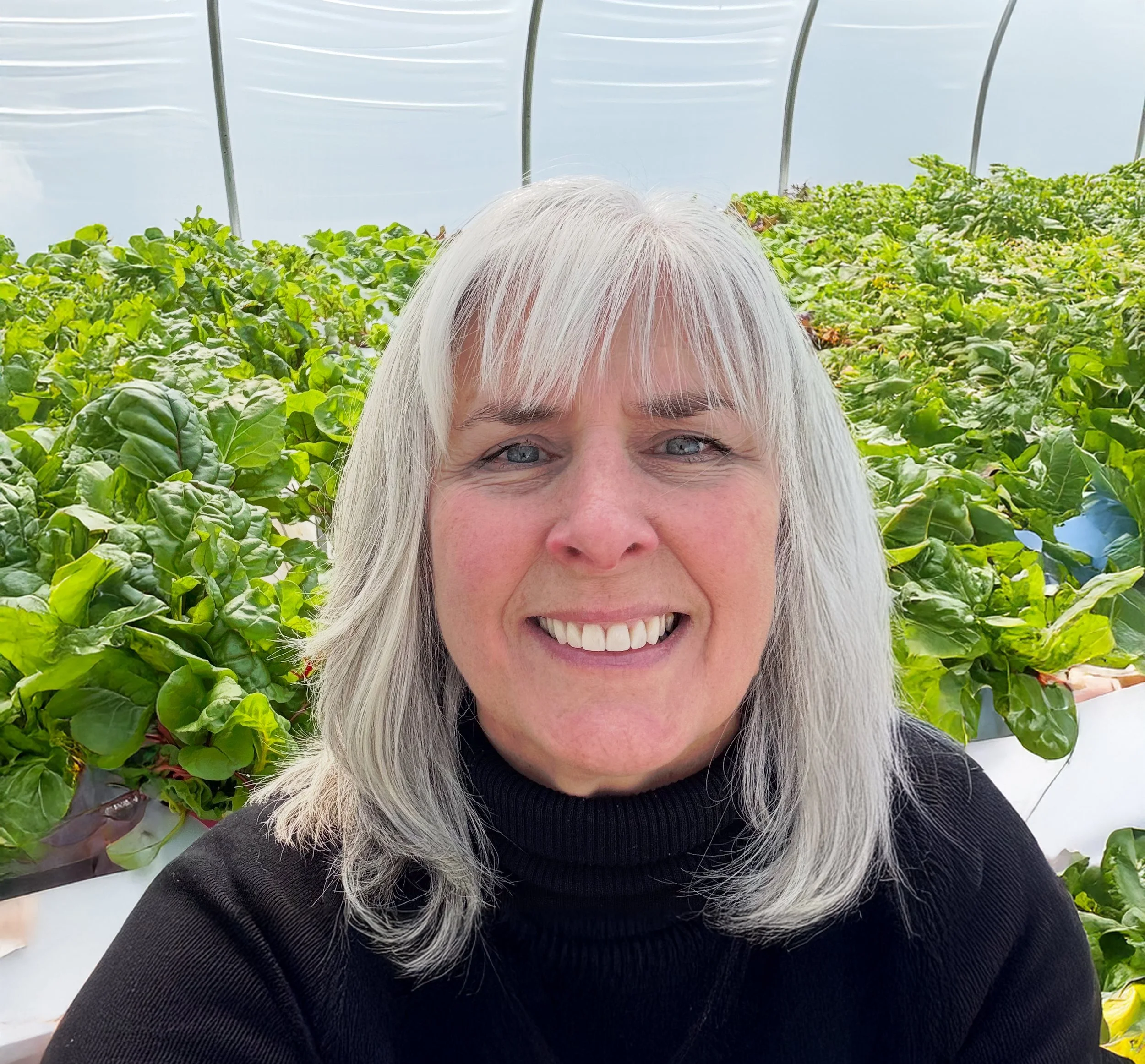 Close-up of a woman with long gray hair smiling in a greenhouse filled with green leafy plants.
