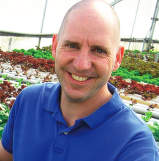 Man smiling in a greenhouse with rows of green and red lettuce plants.