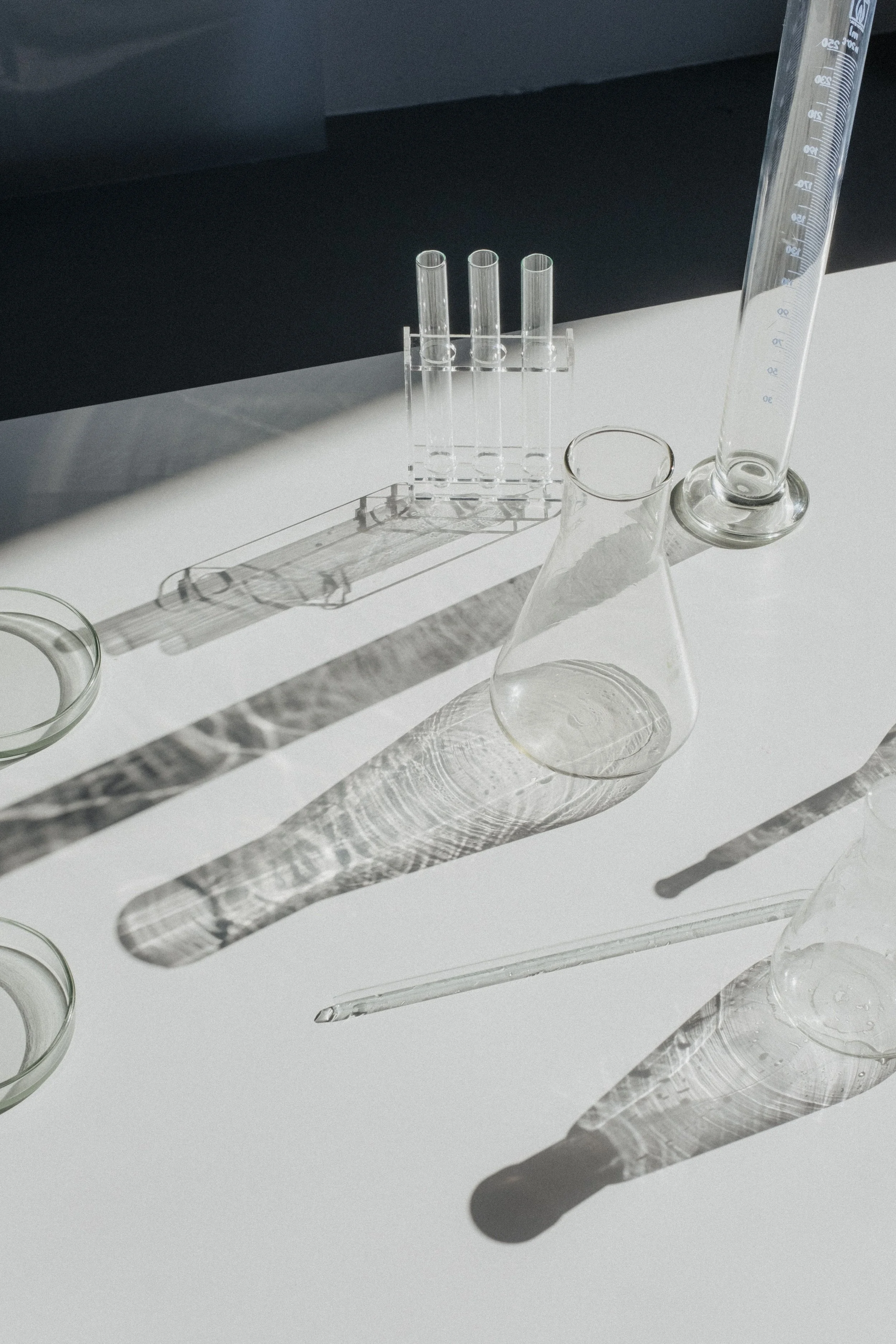 Laboratory glassware including test tubes, a beaker, a graduated cylinder, and a glass stir rod on a white surface, with shadows cast by the glass objects.