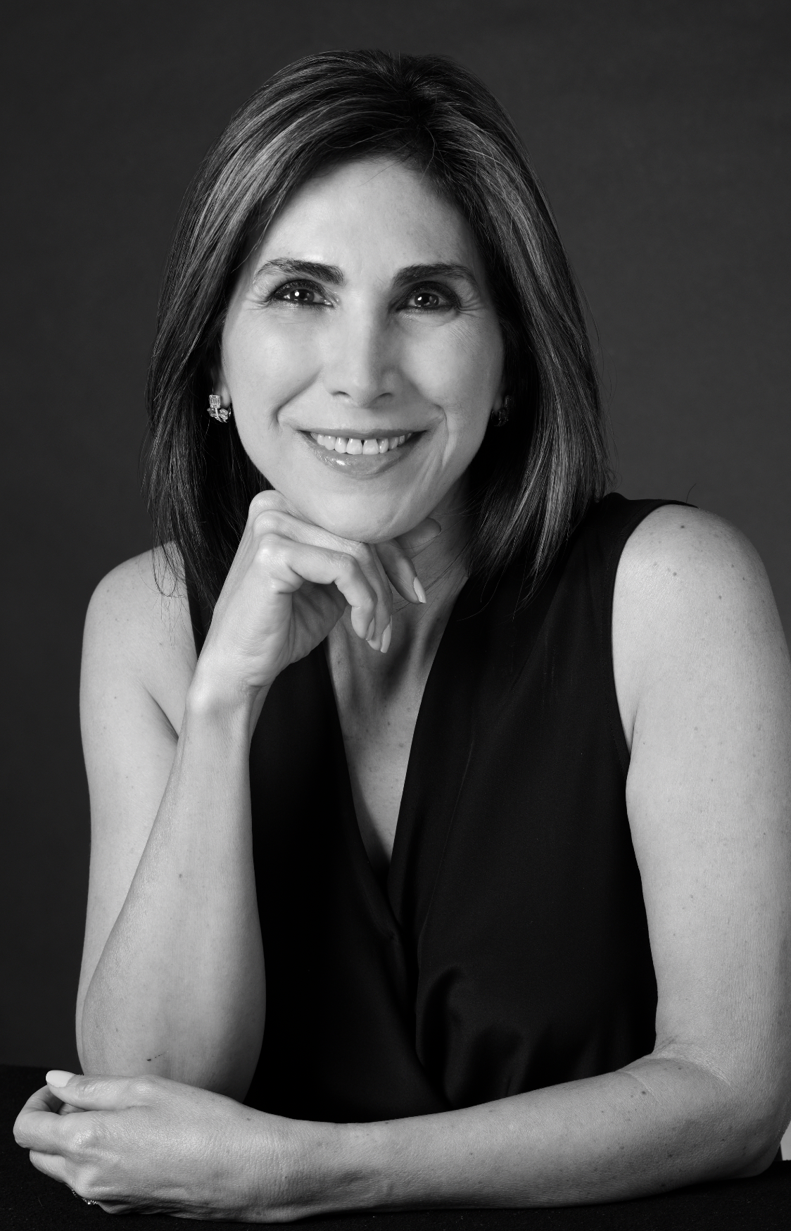 Black and white portrait of a smiling woman with shoulder-length hair, wearing earrings and a sleeveless black top, resting her chin on her hand against a dark background.