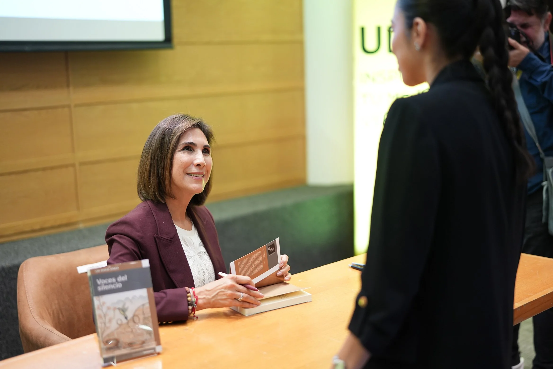 A woman with shoulder-length brown hair, wearing a maroon blazer and holding a book, is seated at a table. She is engaging in conversation with a woman standing in front of her, who has long dark hair tied in a braid and is wearing a black jacket. In the background, a man with dark hair and a beard takes a photograph with a camera. The setting appears to be an indoor event or book signing, with books and informational materials on the table.