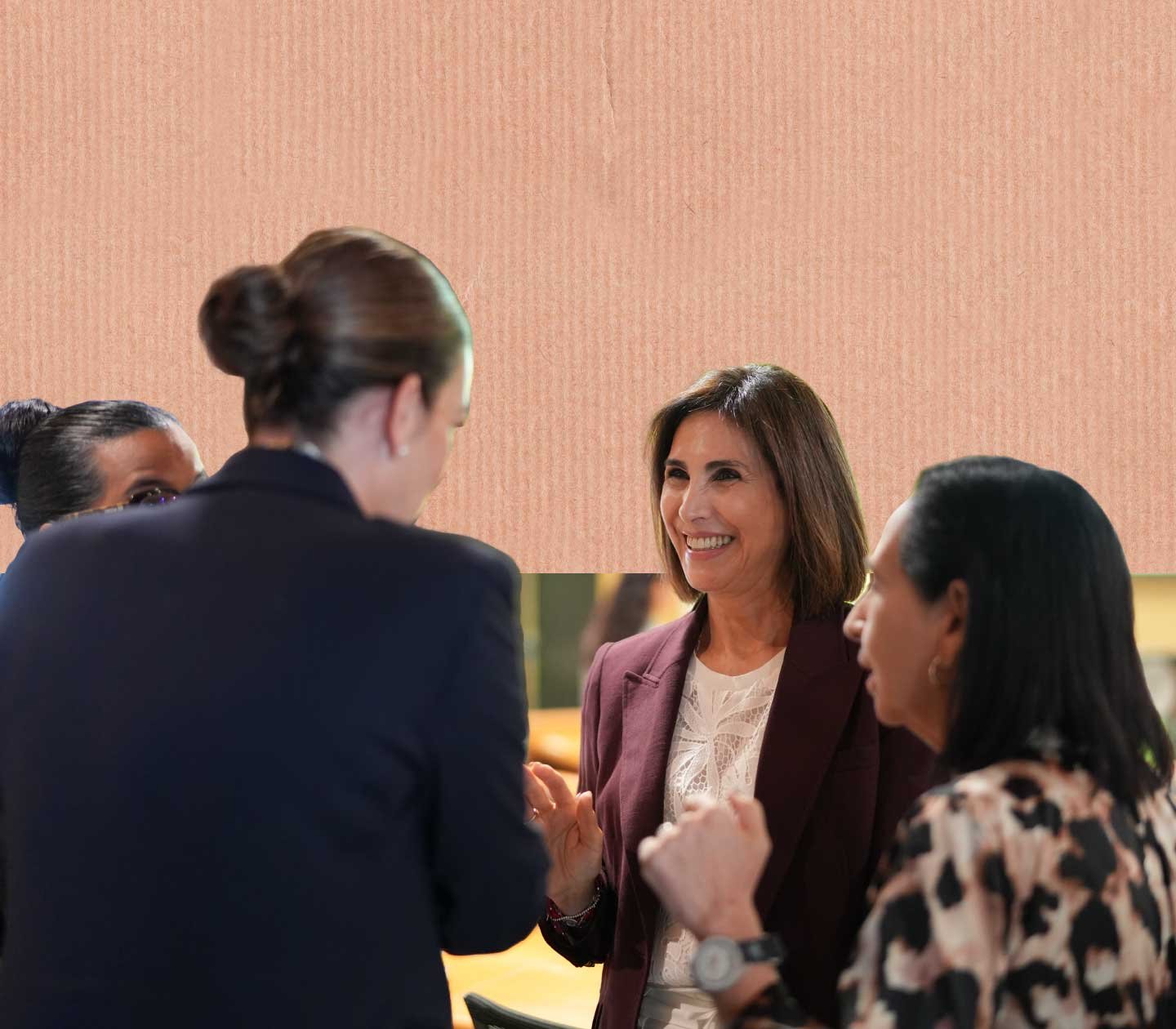 Four women engaged in a conversation, two women visible from the front and two from behind, one smiling and wearing a burgundy blazer and white blouse, the other gesturing with hands, in an indoor setting.