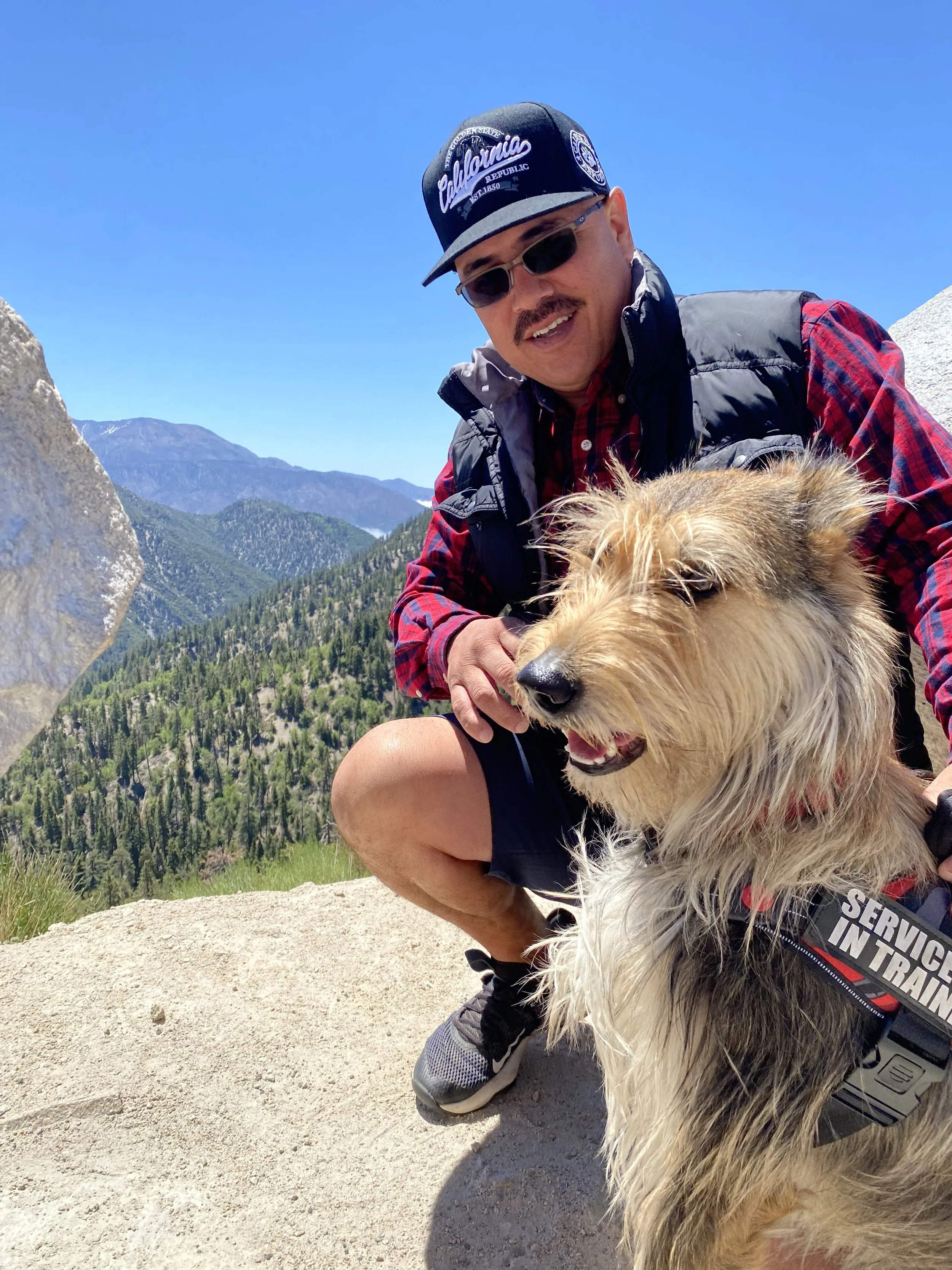 A man in outdoor gear and sunglasses crouches with a large golden retriever dog in a mountainous outdoor setting under a bright blue sky.