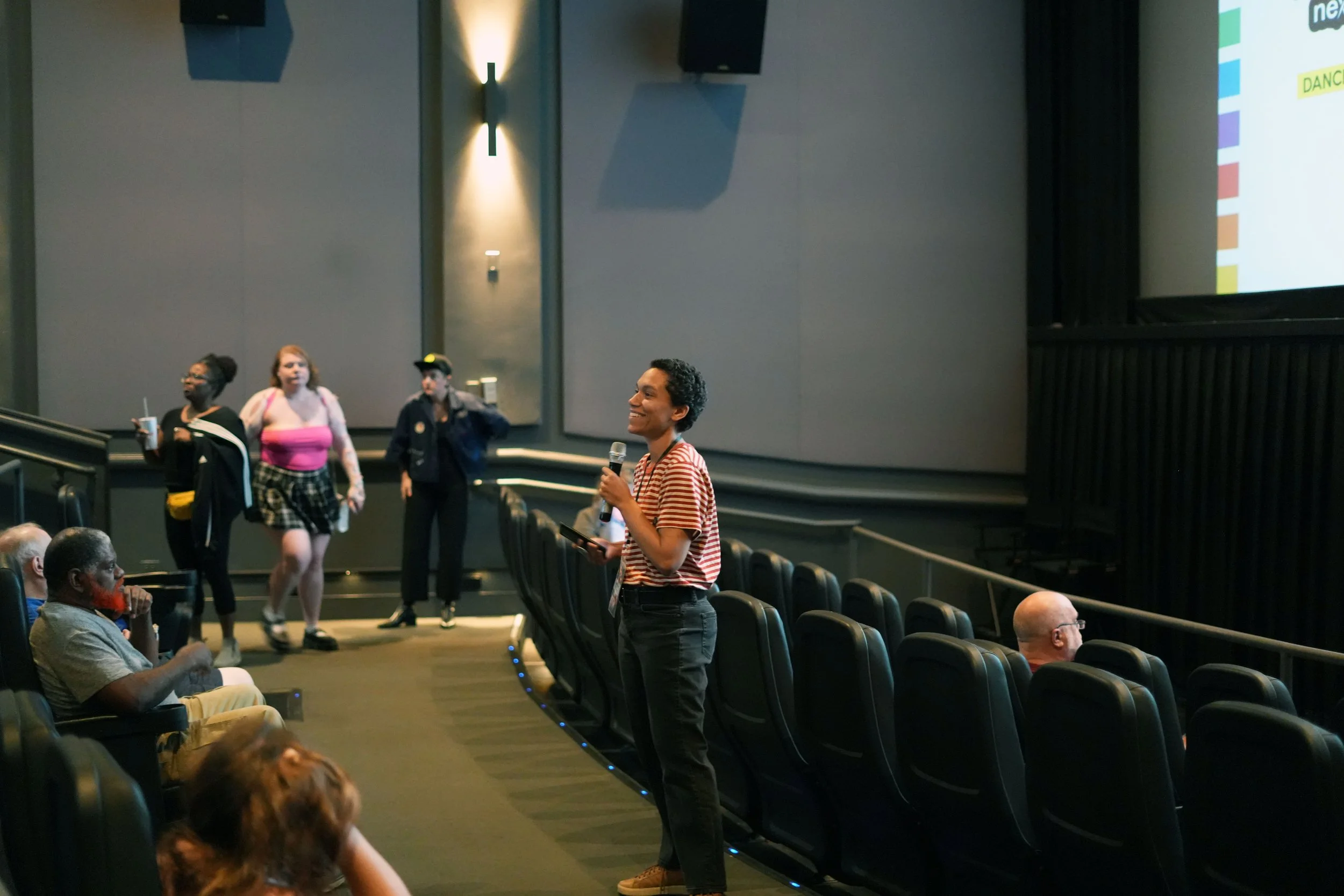 A woman holding a microphone stands in an auditorium with several empty seats, smiling and engaging with an audience. Behind her, a group of people walks across the stage, some with bags and drinks. The auditorium has dim lighting, with a large screen on the right side displaying colorful content.