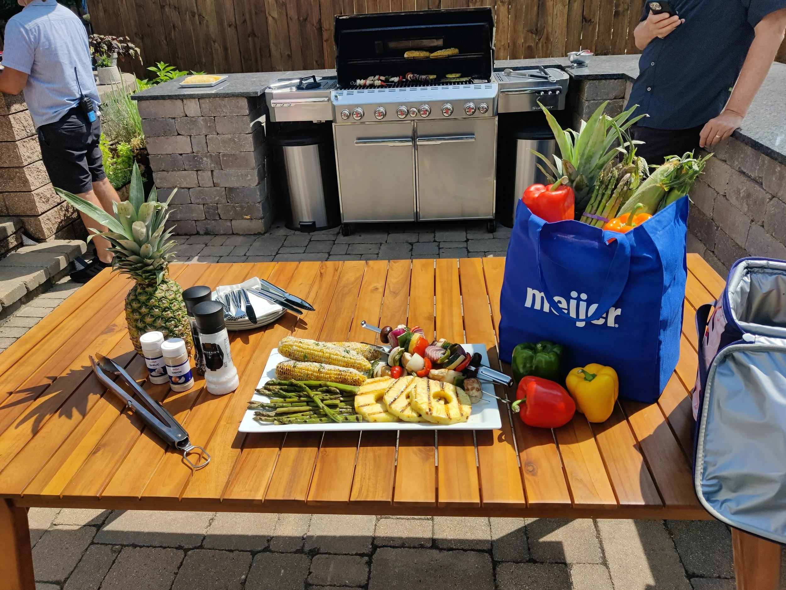 A wooden outdoor dining table set with food including grilled vegetables, corn on the cob, pineapple, and peppers, with utensils and condiments. In the background, a man is grilling on a barbecue grill, and there is a blue reusable grocery bag with asparagus, peppers, and pineapple outside.