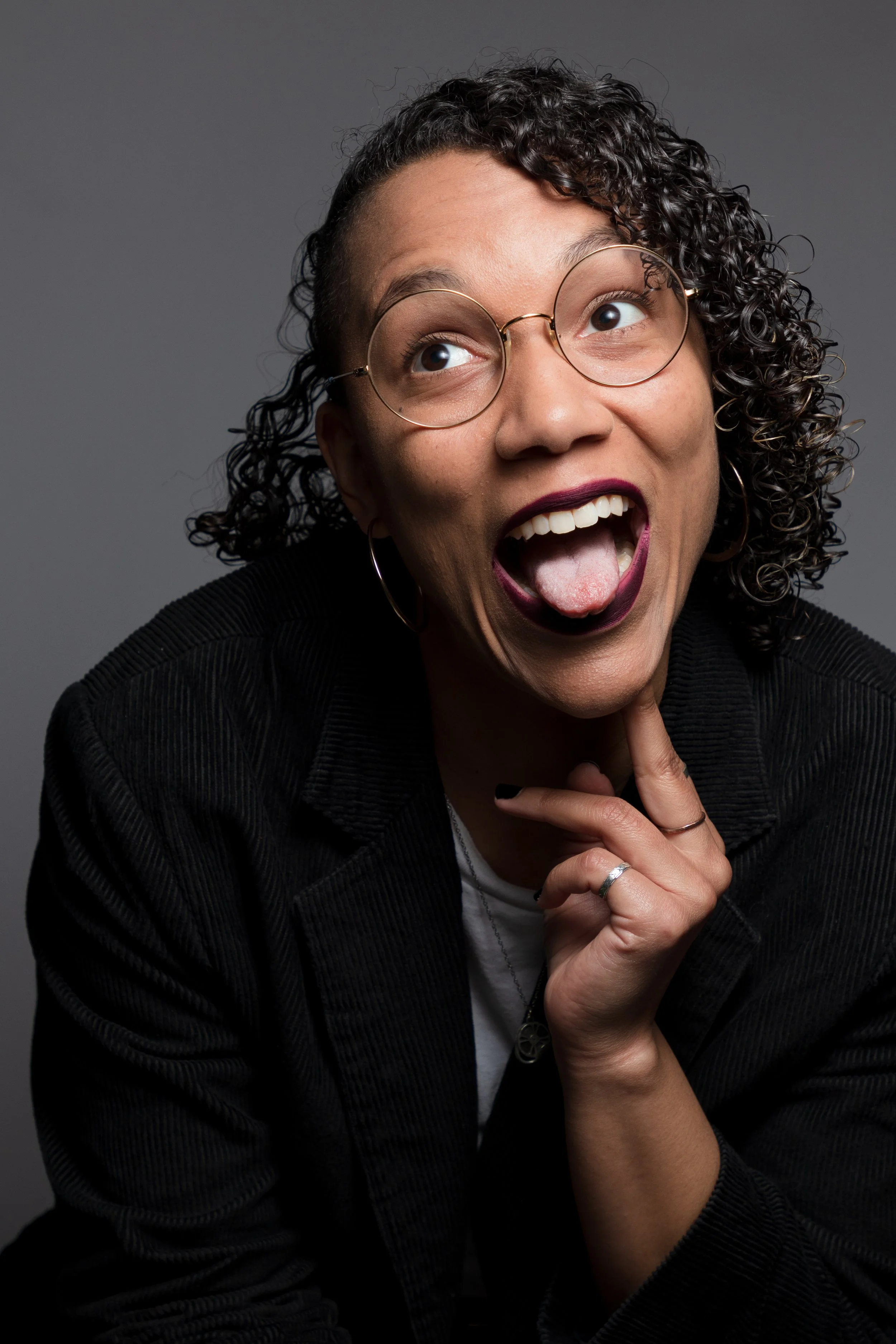 A woman with curly hair and glasses making a playful face with her tongue out and eyes looking upward, against a dark background.