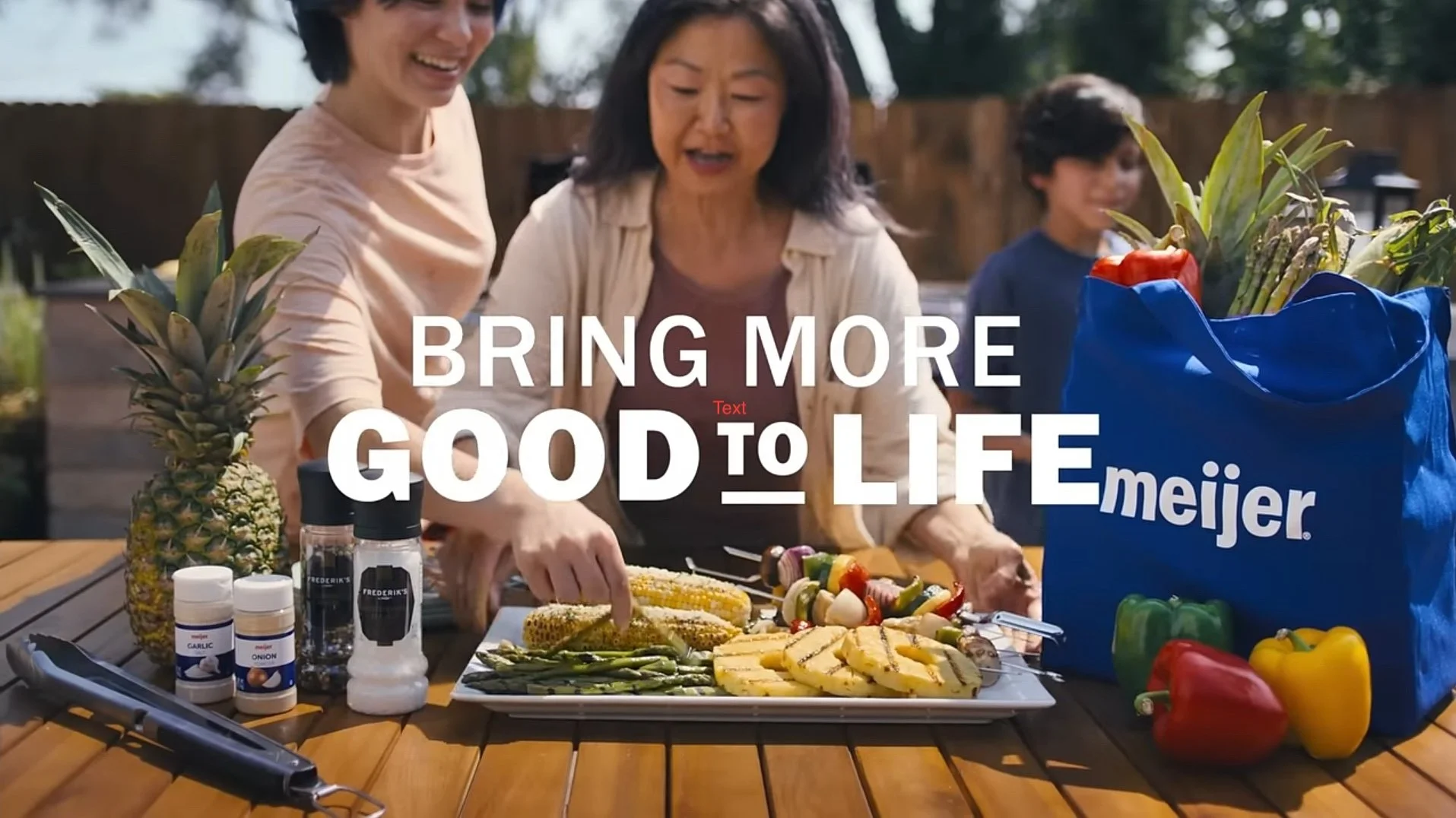 Three women enjoy a outdoor meal with vegetables, pineapple, and grilled corn on a table, with a large blue Meijer store shopping bag among the food. Overlaid text reads, 'Bring More Good to Life.'