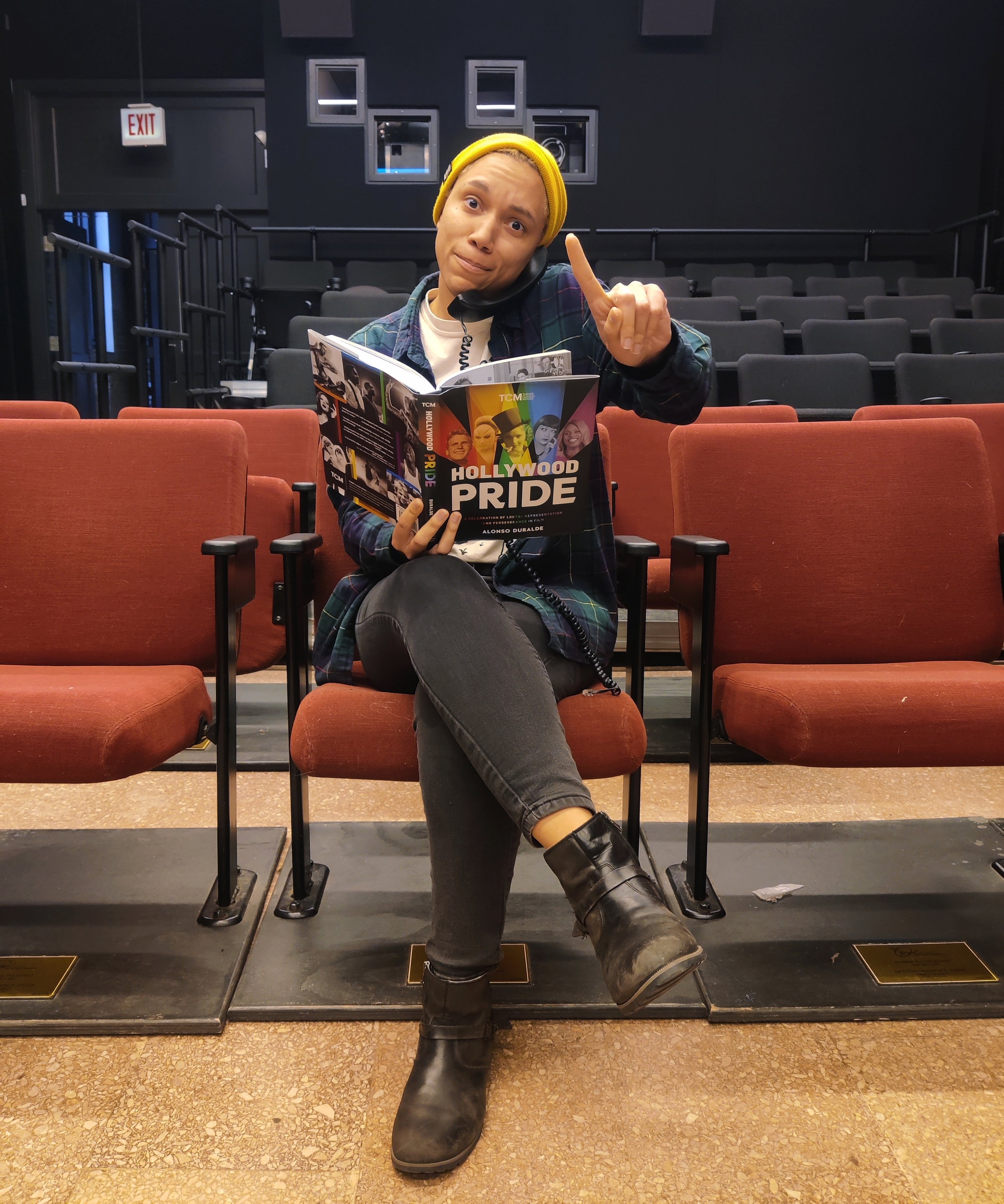 A person sitting in an auditorium with blank chairs, wearing a yellow beanie, holding a 'Hollywood Pride' magazine, talking on a phone, with one leg crossed over the other, and making a gesture with one finger, surrounded by empty seats and a dark background.