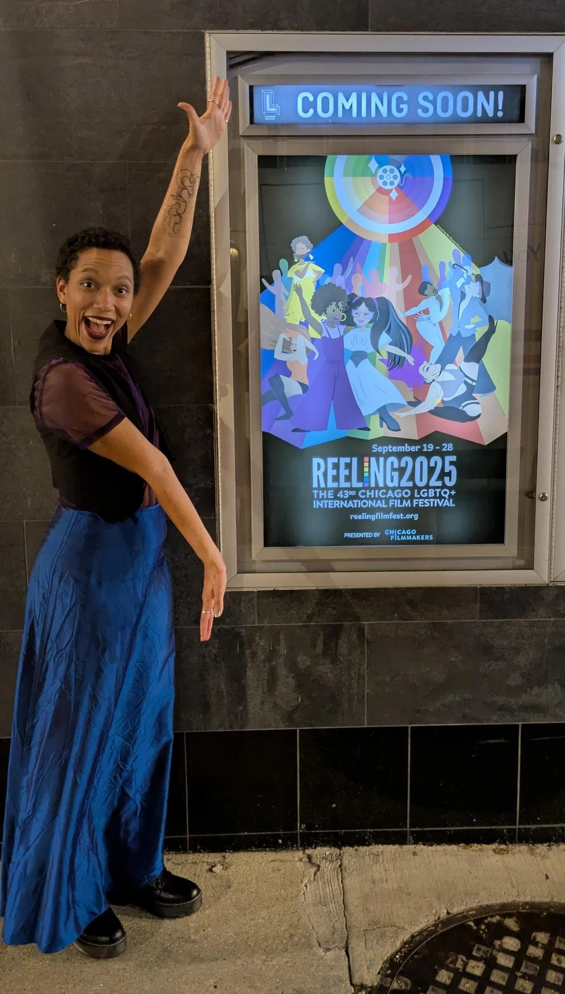 A woman with short curly hair, tattooed arm, and dressed in a black top with sheer sleeves and a long blue skirt, pointing to a digital poster for the Chicago LGBTQ+ International Film Festival Reeling 2025, scheduled from September 19 to 28. The poster features colorful animated illustrations of diverse women and a blue circle with a rainbow pattern at the top.