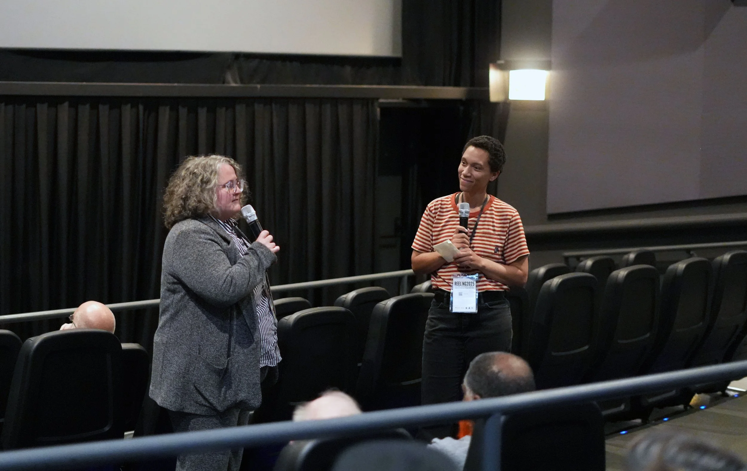 Two women are standing in an auditorium, holding microphones and engaging in conversation. The woman on the left has curly blonde hair and glasses, wearing a grey blazer, while the woman on the right has short dark hair and is dressed in a striped t-shirt, wearing a name badge that says 'REELING 2025'. The auditorium has black chairs arranged in rows, and a few people are seated in the background.