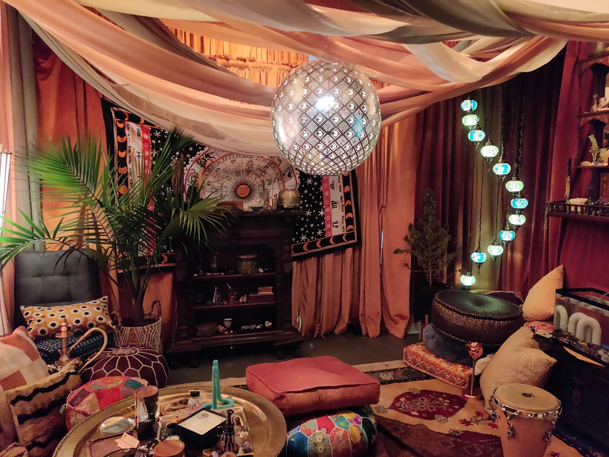 A bohemian-style living room decorated with draped fabric on the ceiling, a large hanging geometric lamp, and a tapestry of astrologic symbols on the wall. The space includes a variety of cushions, plants, and eclectic furniture with colorful textiles and string lights.